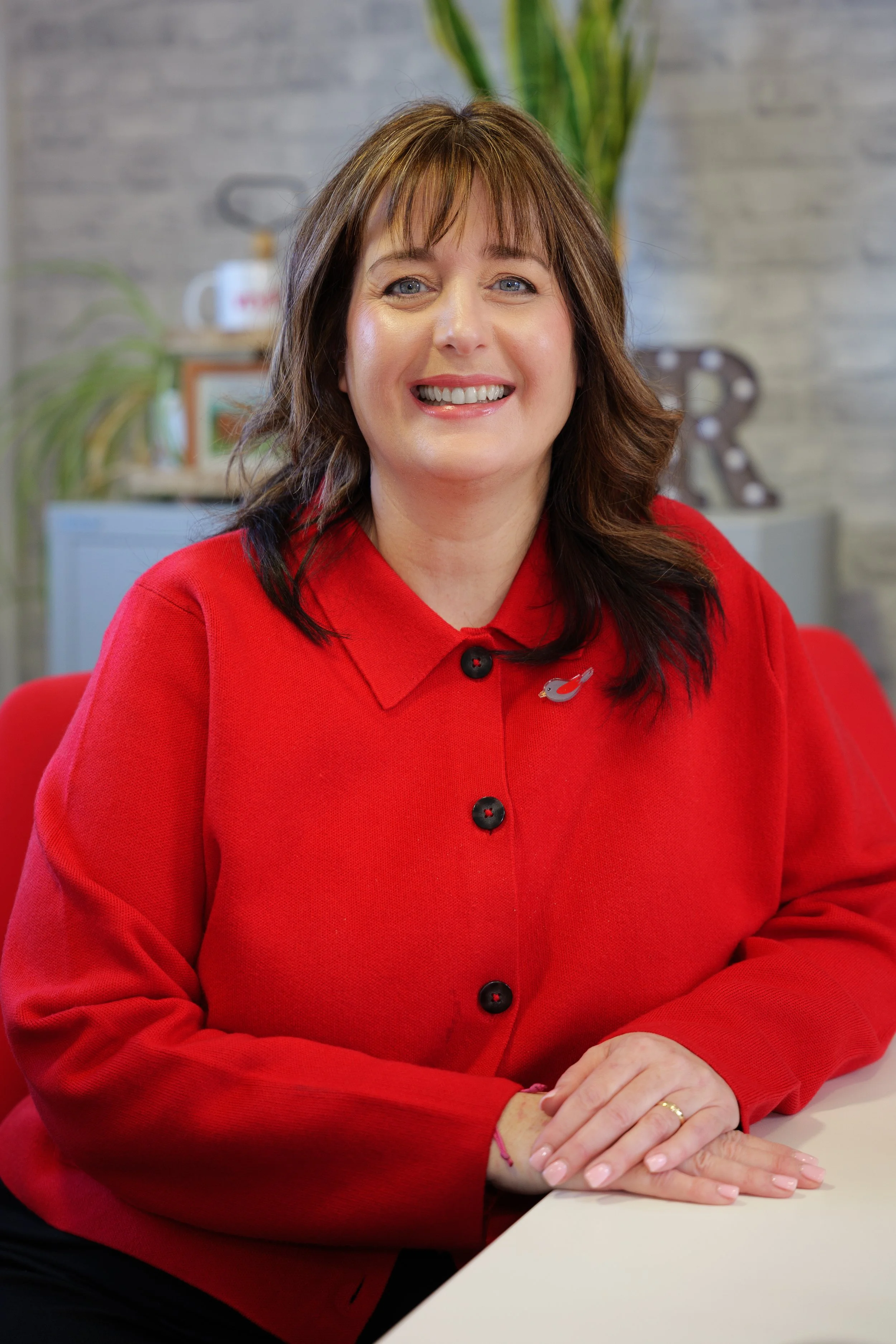 A woman with shoulder-length brown hair, wearing a red button-up shirt, smiling while sitting at a table with her hands resting on it. The background shows a plant and a decorative letter 'R' with lights.