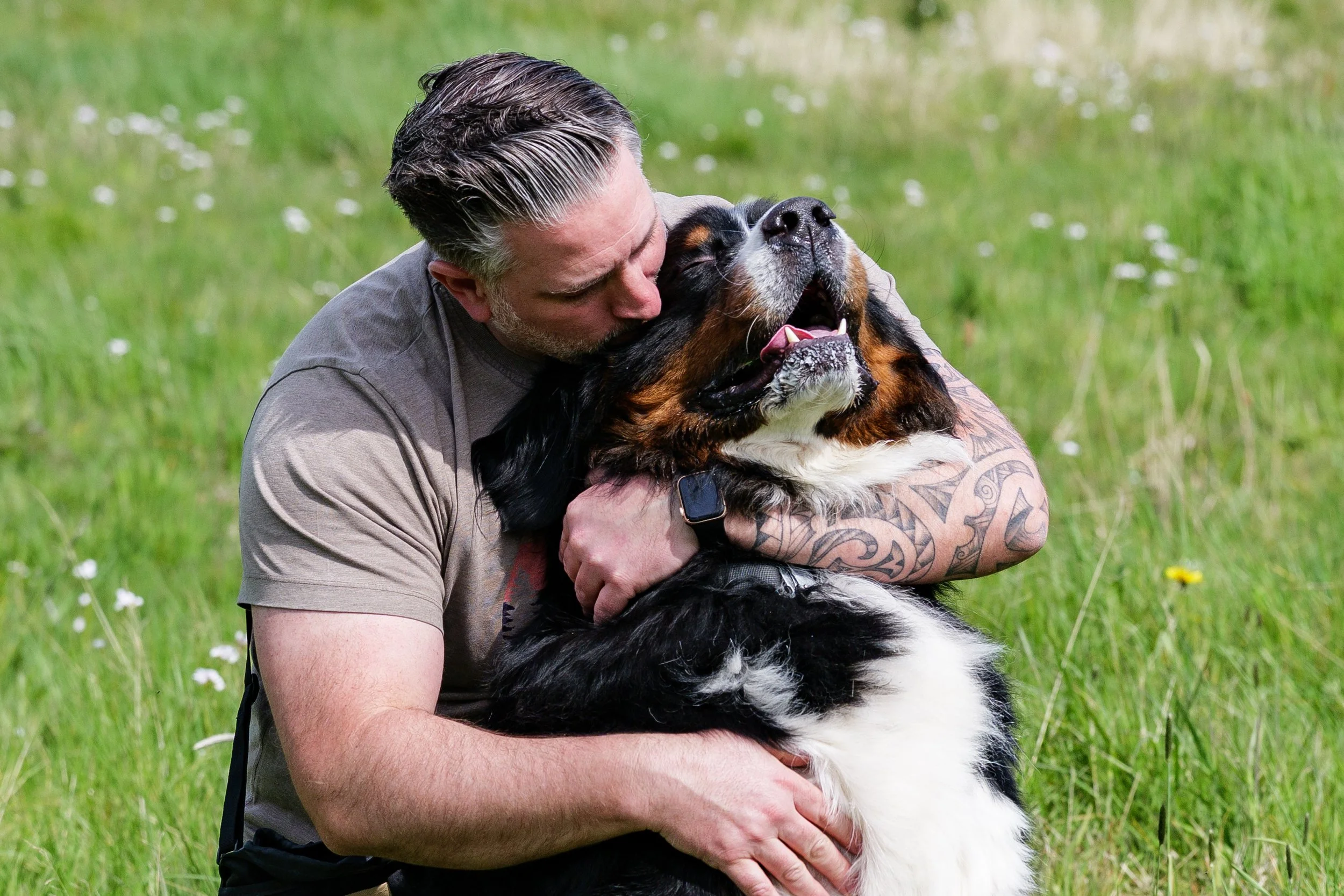 A man with tattoos hugging a large Bernese Mountain Dog outdoors on a grassy field.