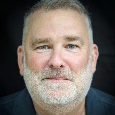 Close-up portrait of a middle-aged man with gray hair and beard, blue eyes, and a neutral expression.