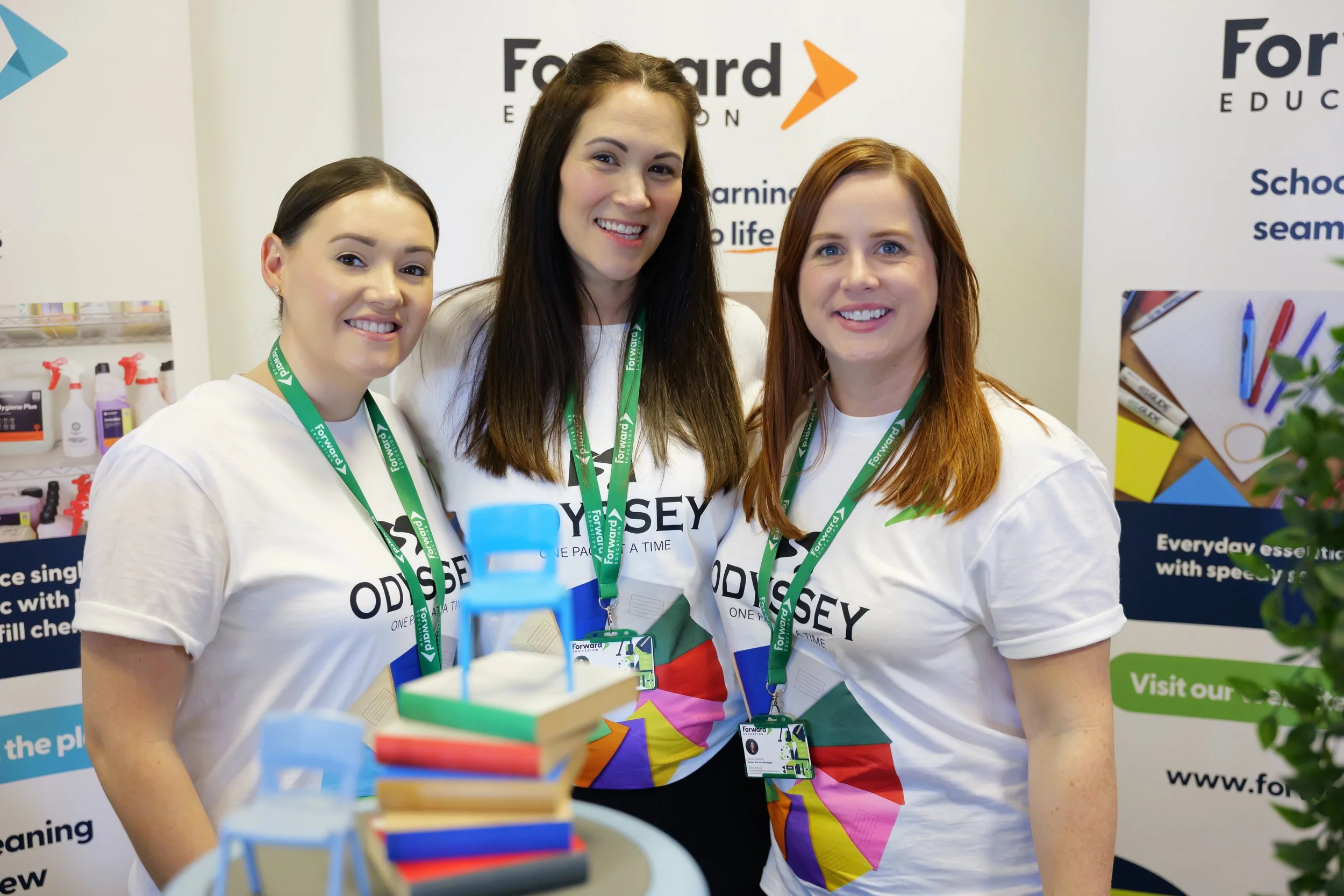 Group of three women smiling at an educational event, wearing white t-shirts with 'Odyssey' printed on them, and green lanyards with 'Forward' written on them, behind an educational booth with posters, books, and educational materials.