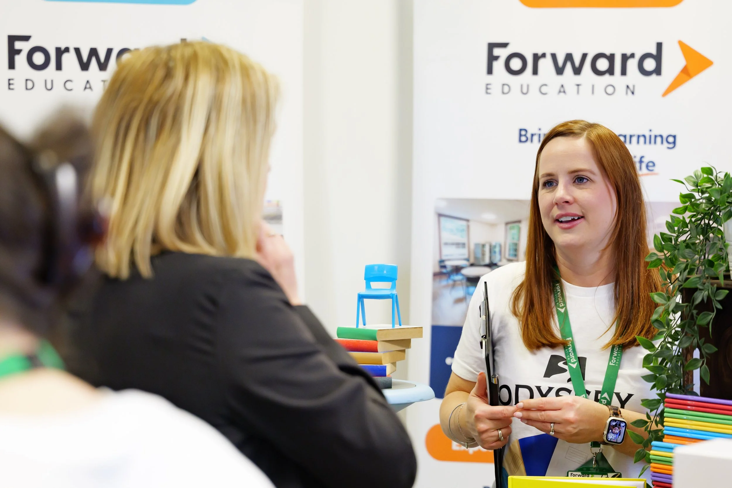 A woman with red hair and a white t-shirt talking to a woman with blonde hair at an education booth with a logo that says 'Forward Education'.