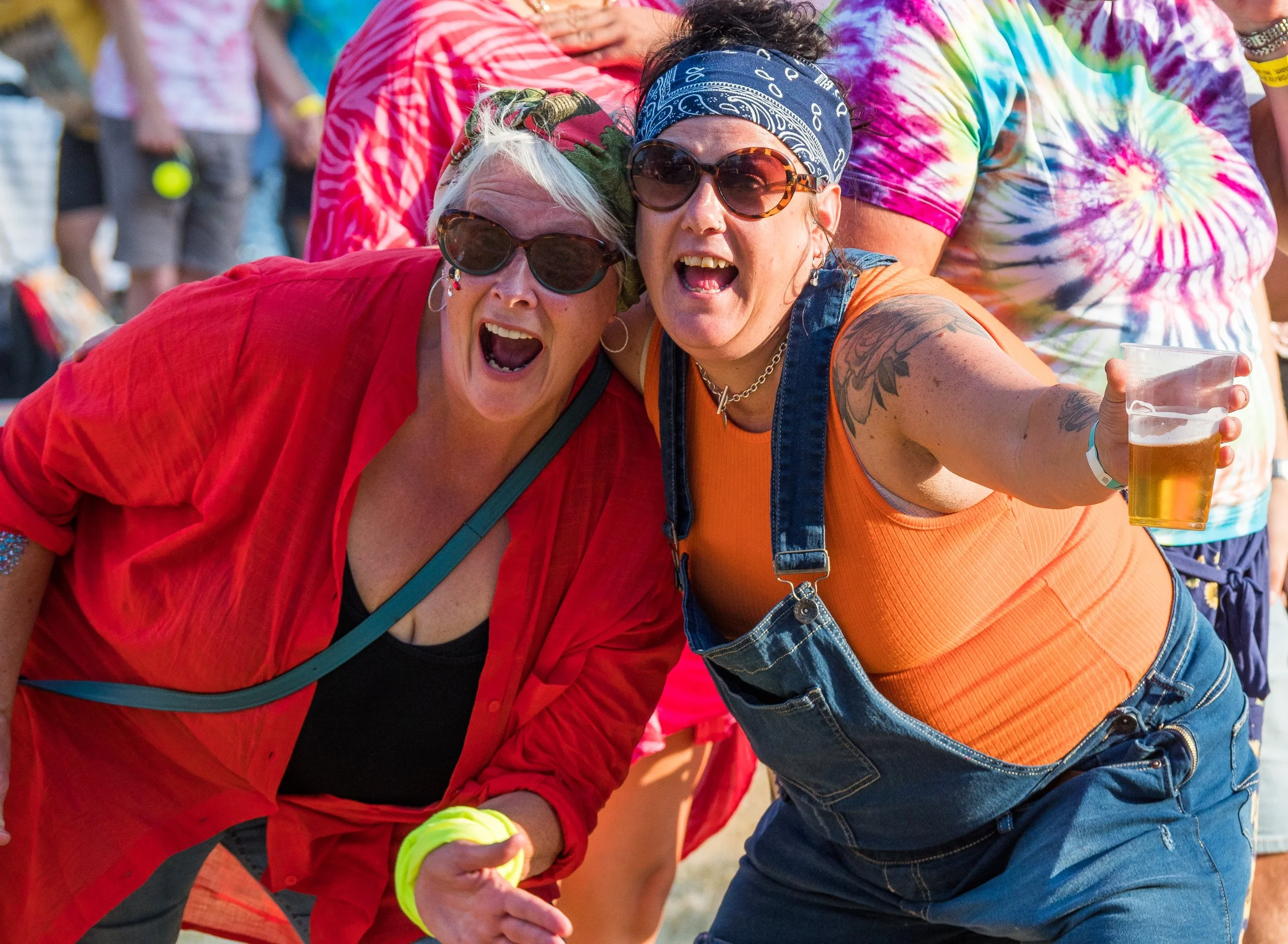 Two women wearing sunglasses and bandanas, at a casual outdoor event, are holding a yellow balloon, with others in colorful tie-dye shirts in the background.