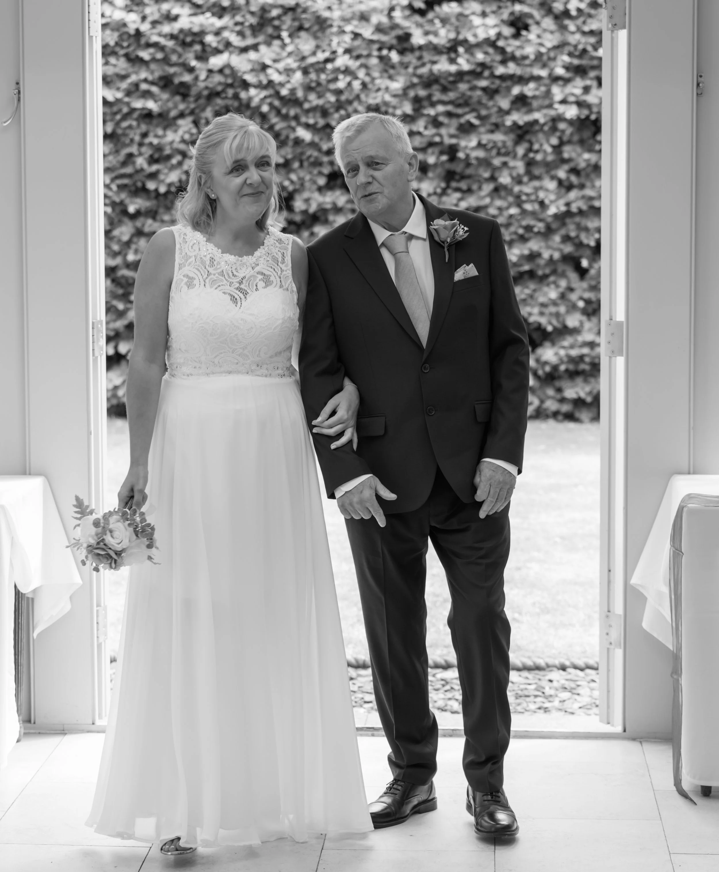 A bride and an older man, possibly her father, are standing at the entrance of a building outdoors. The bride wears a white lace wedding dress and mosaic jewelry, holding a small bouquet of flowers. The man is dressed in a dark suit with a boutonnier