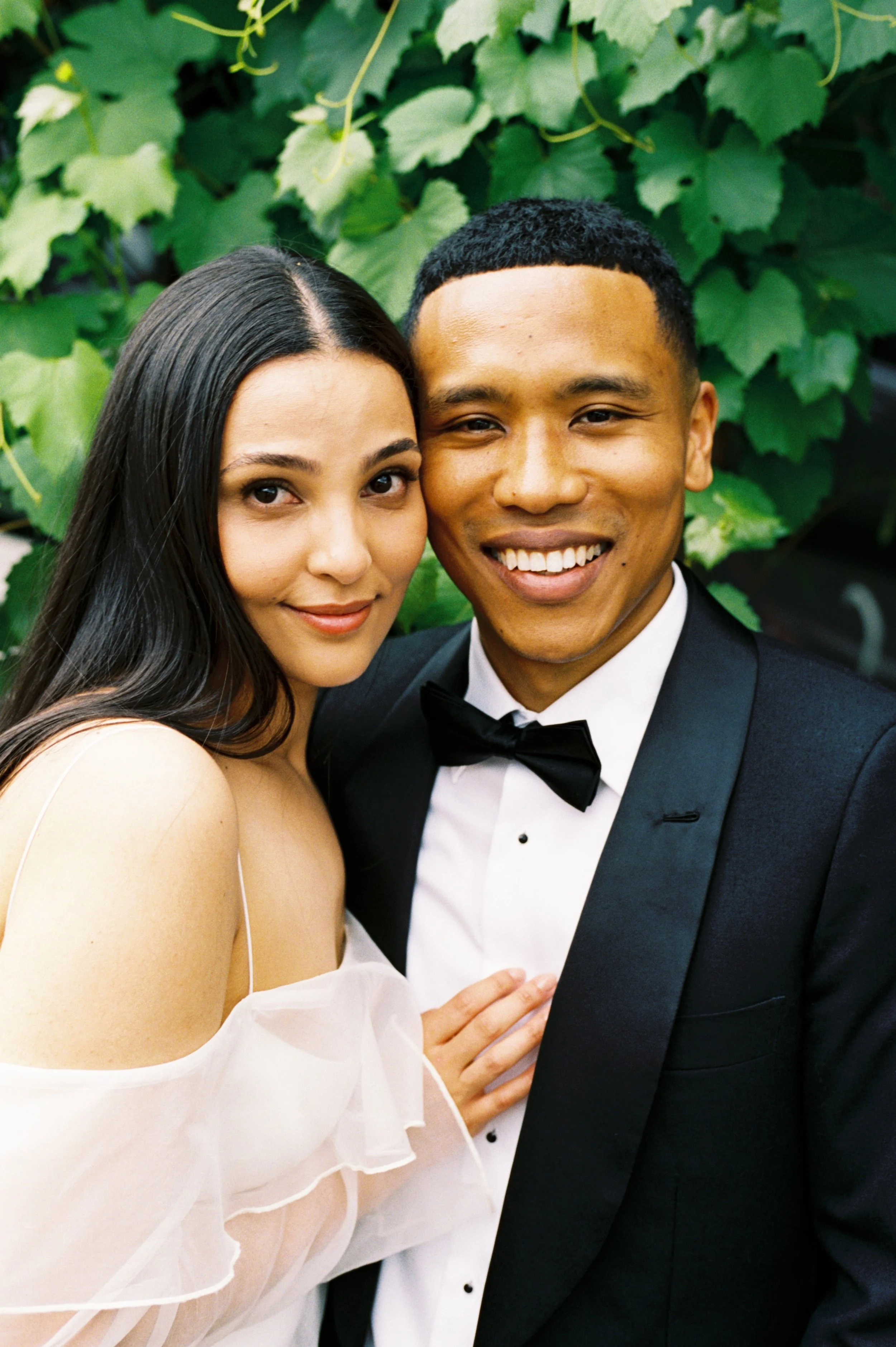 A smiling couple dressed in formal attire, standing close together in front of green leafy plants.
