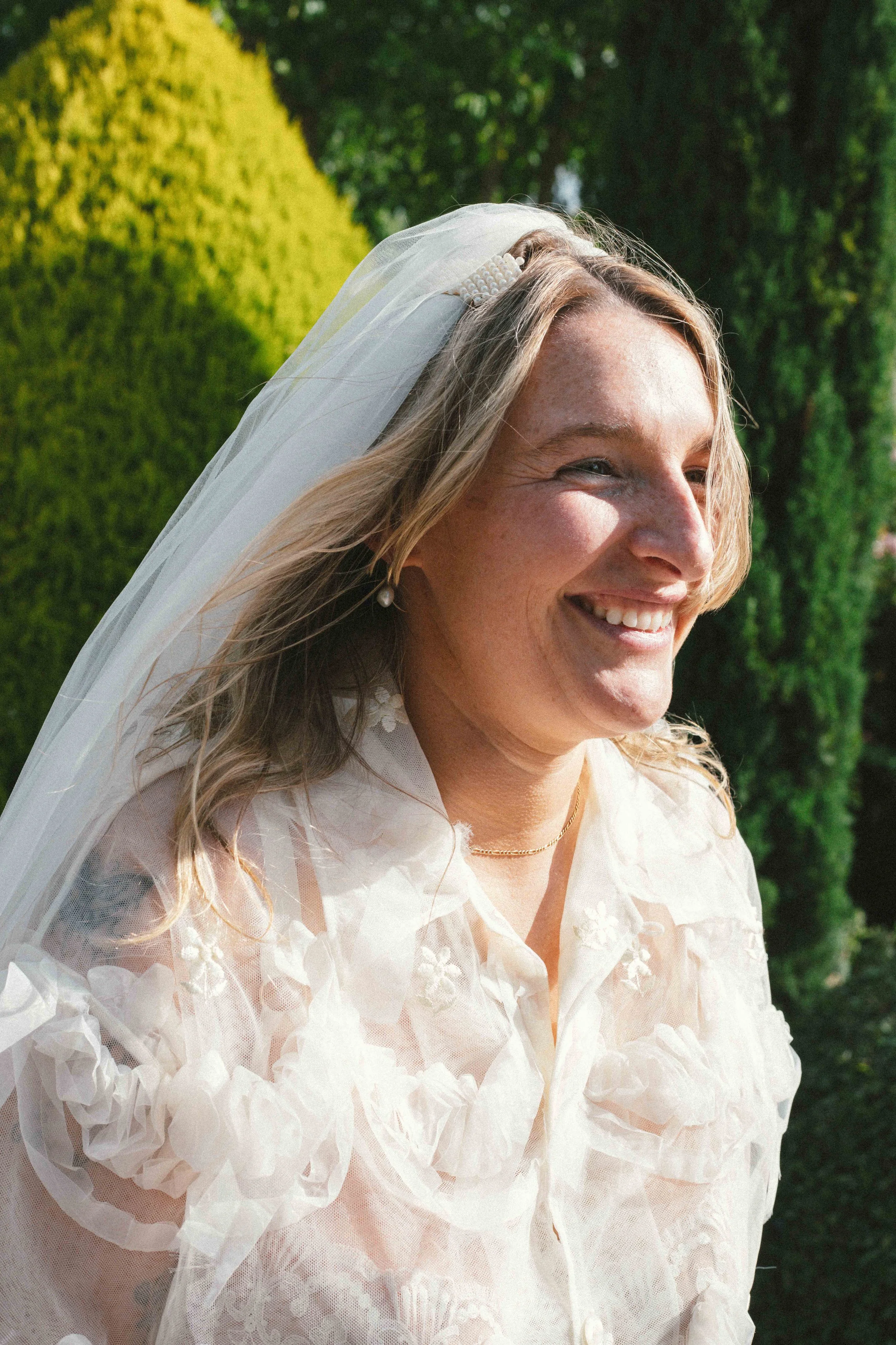 Smiling woman wearing a wedding dress and veil outdoors with green bushes in the background.