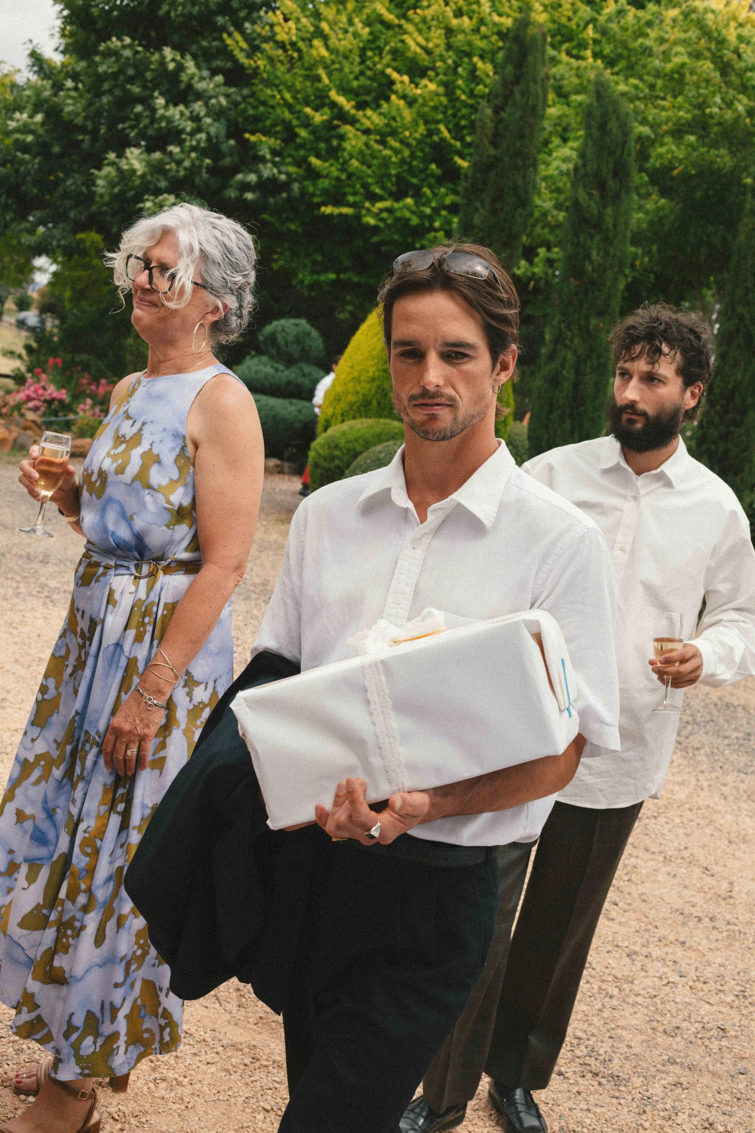 A group of three people at an outdoor event, with a woman holding a glass of champagne and two men carrying gifts, against a backdrop of green trees and bushes.