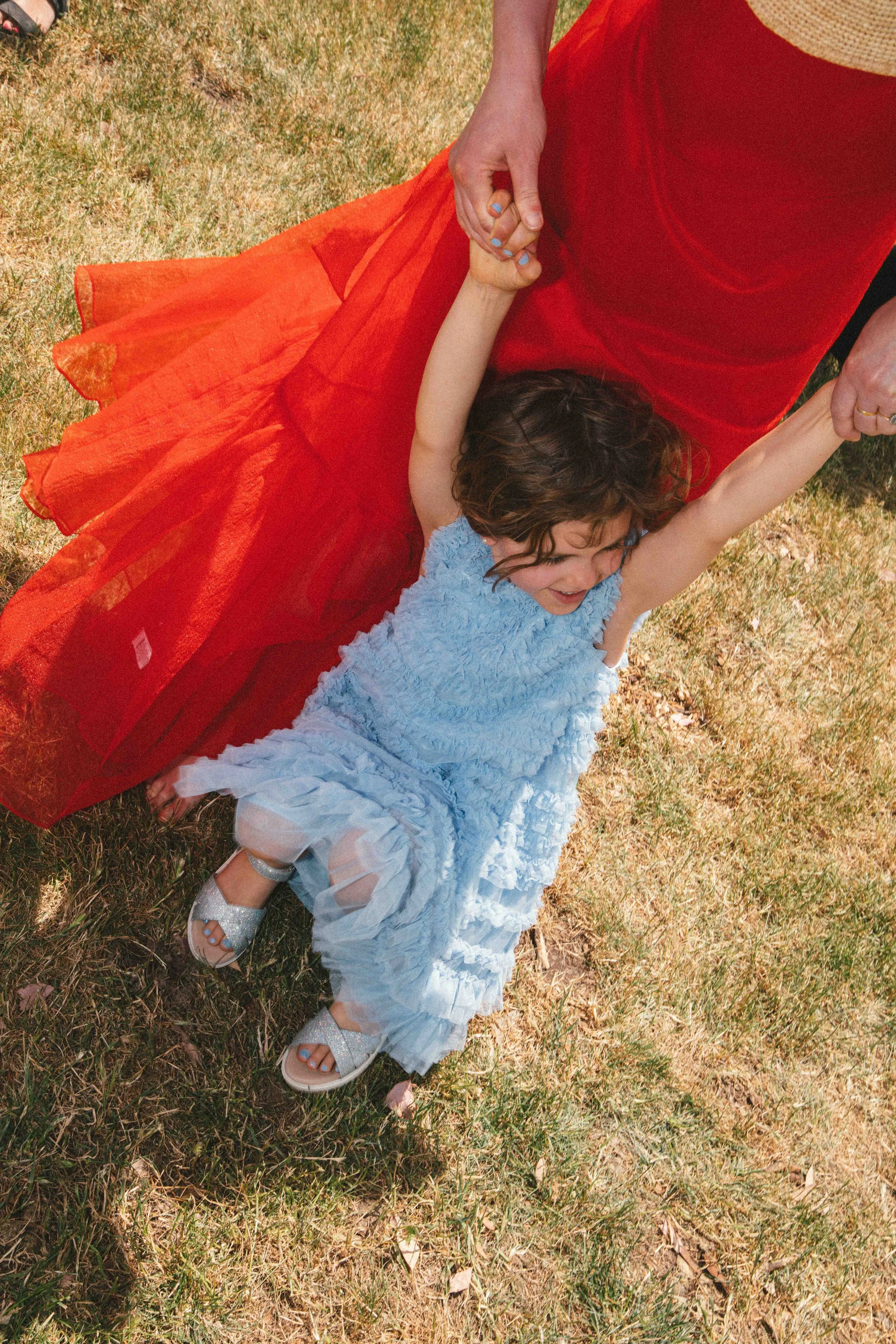 A young girl in a blue dress is being swung around by an adult, holding her hands, outdoors on a grassy area. The girl is smiling with joy.