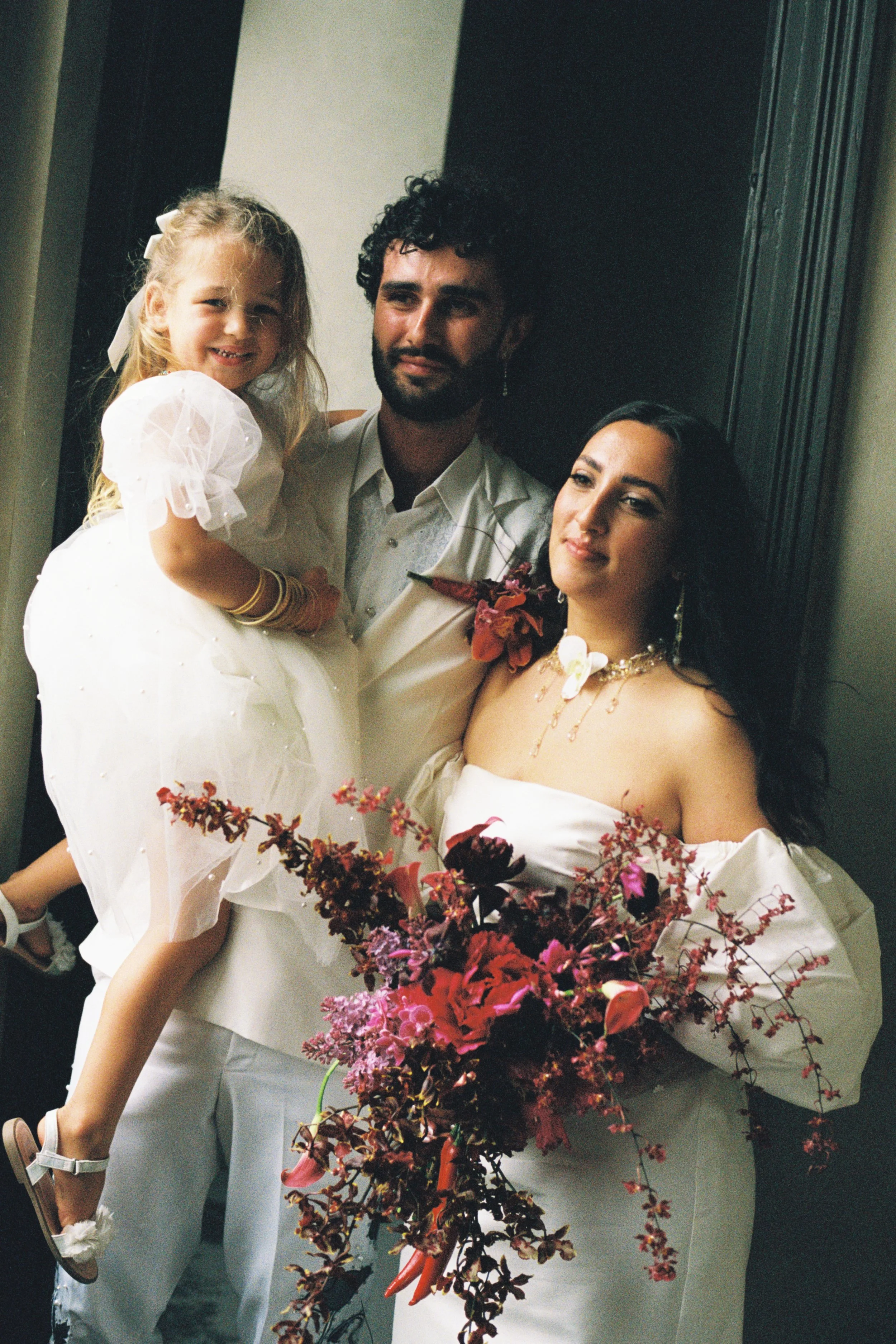 A couple holding a young girl with a bouquet of pink and red flowers, all dressed in white, in an indoor setting.