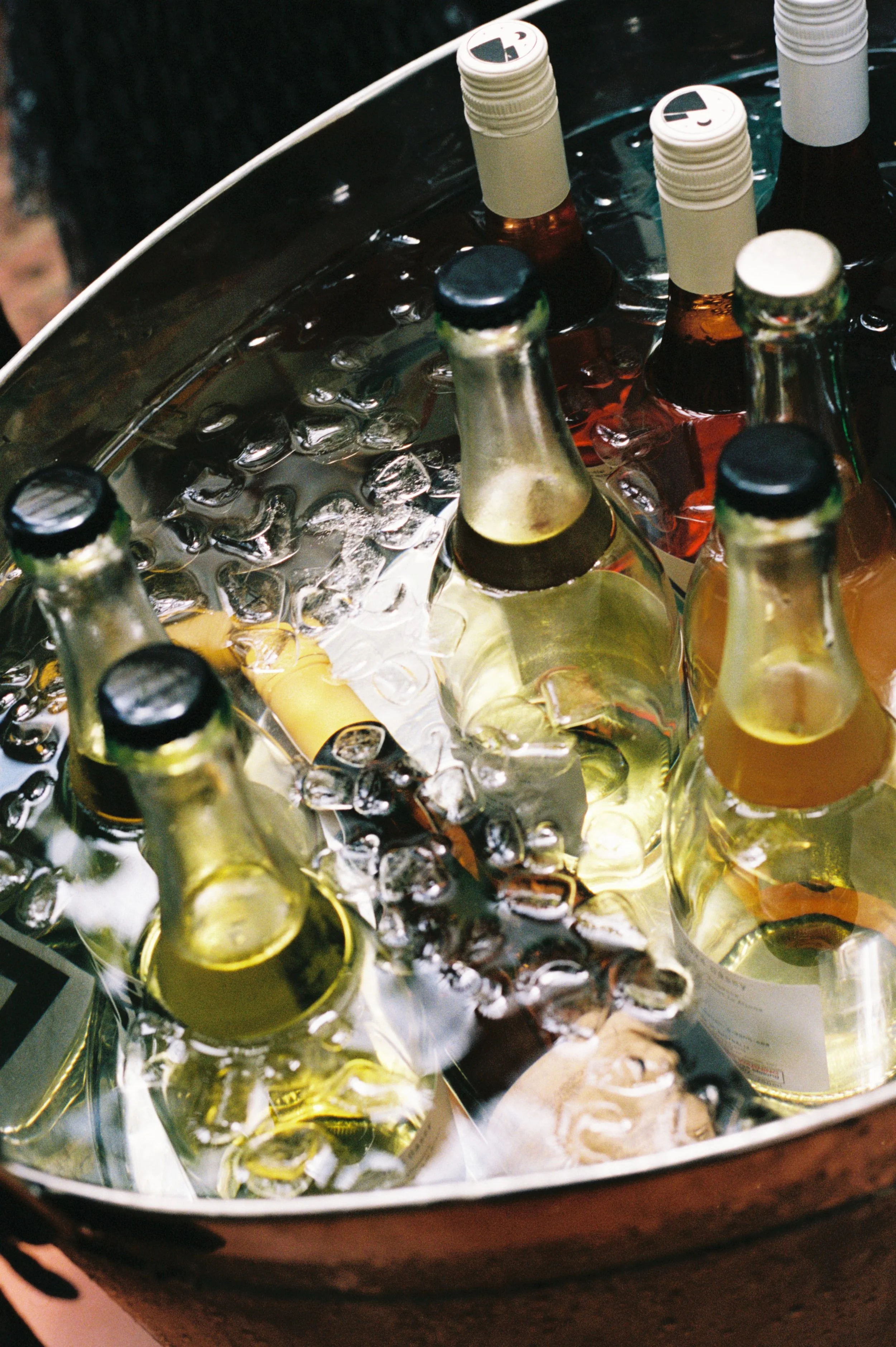 Several bottles of beer and soda chilling in a round metallic ice bucket filled with ice.