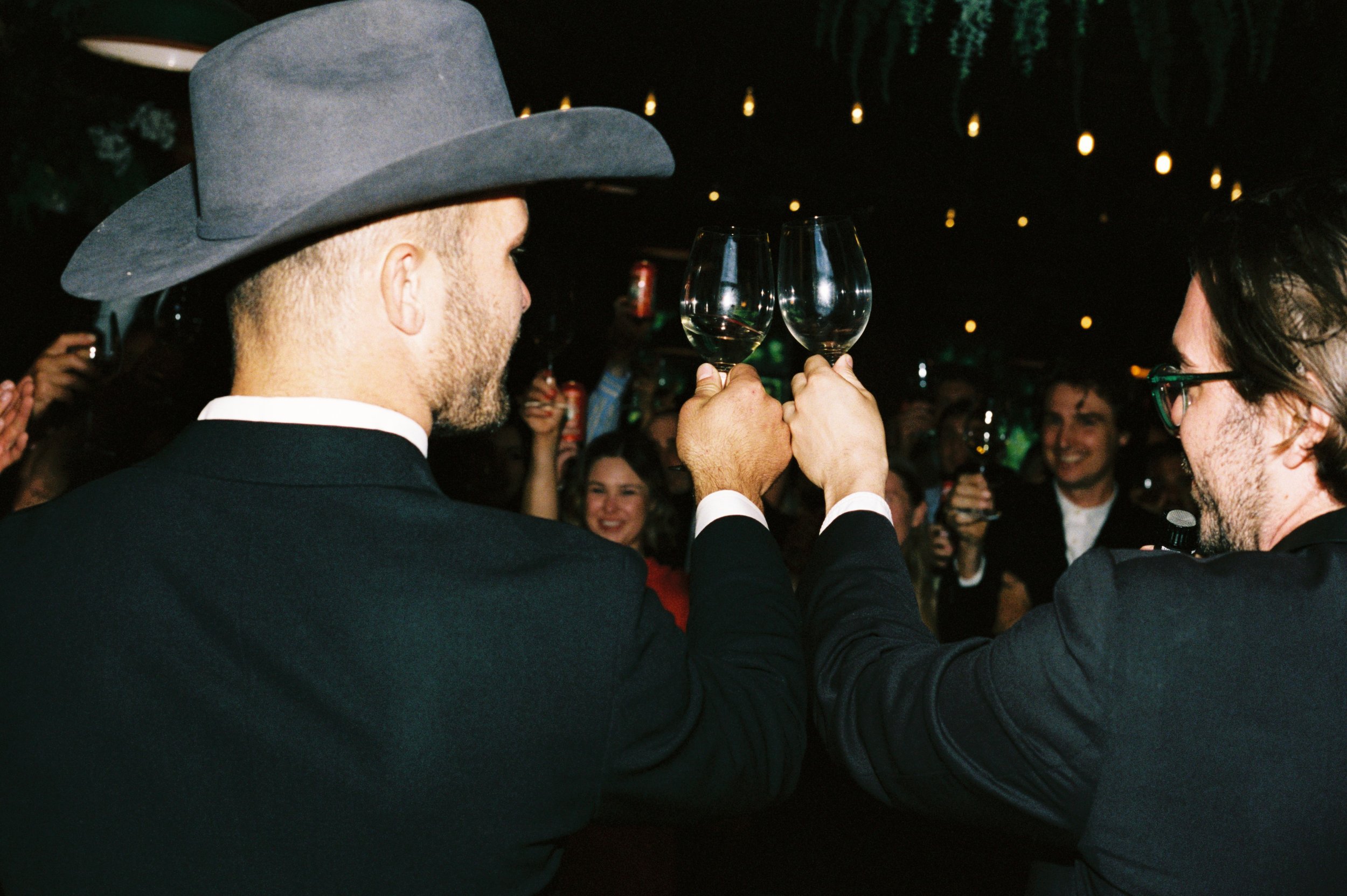 Two men dressed in black tuxedos and one wearing a gray cowboy hat, raising empty wine glasses in a toast at a celebration or party. People in the background are smiling and taking photos.