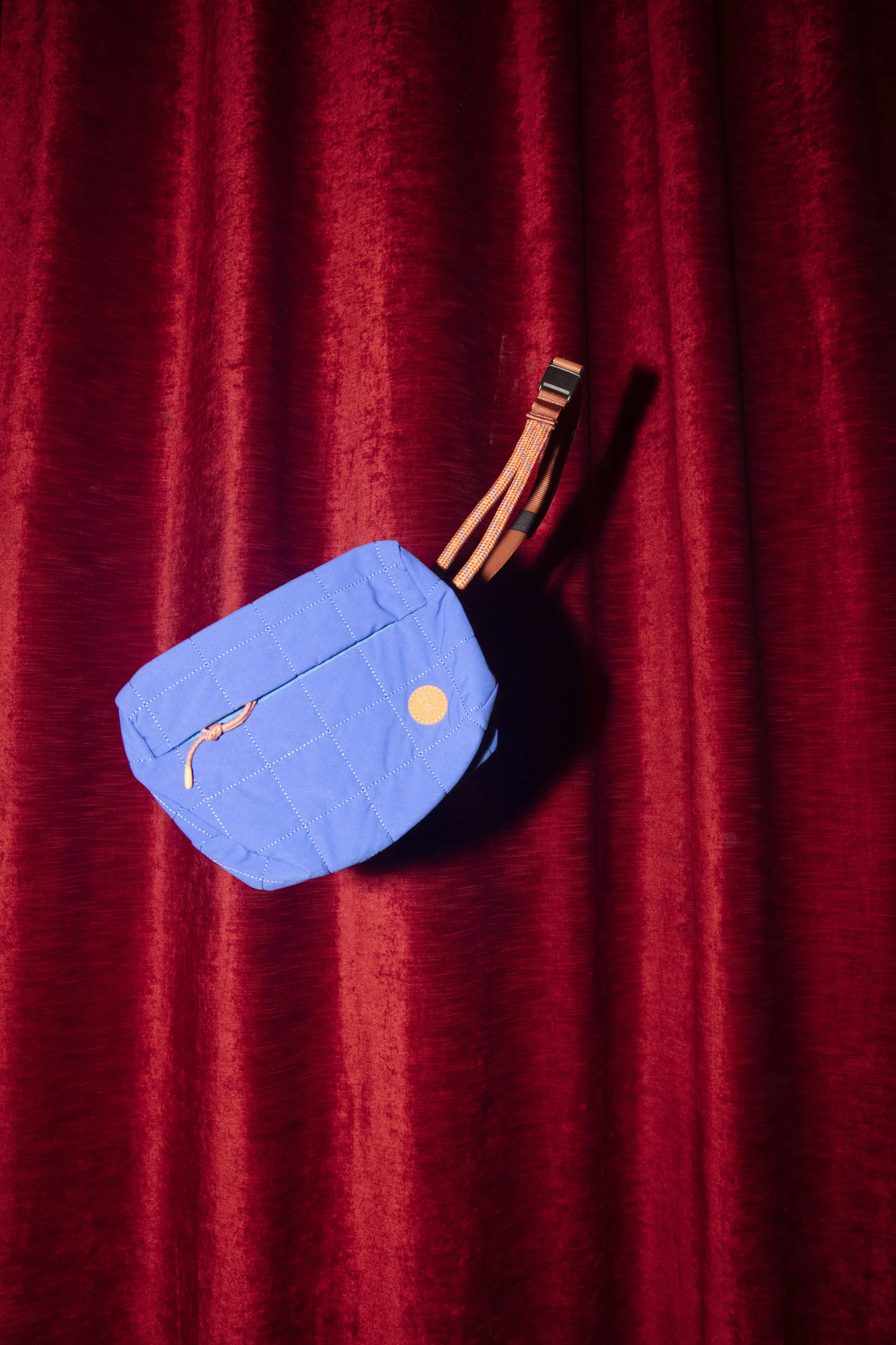 A blue quilted bag with orange drawstrings hanging on a red velvet curtain background.