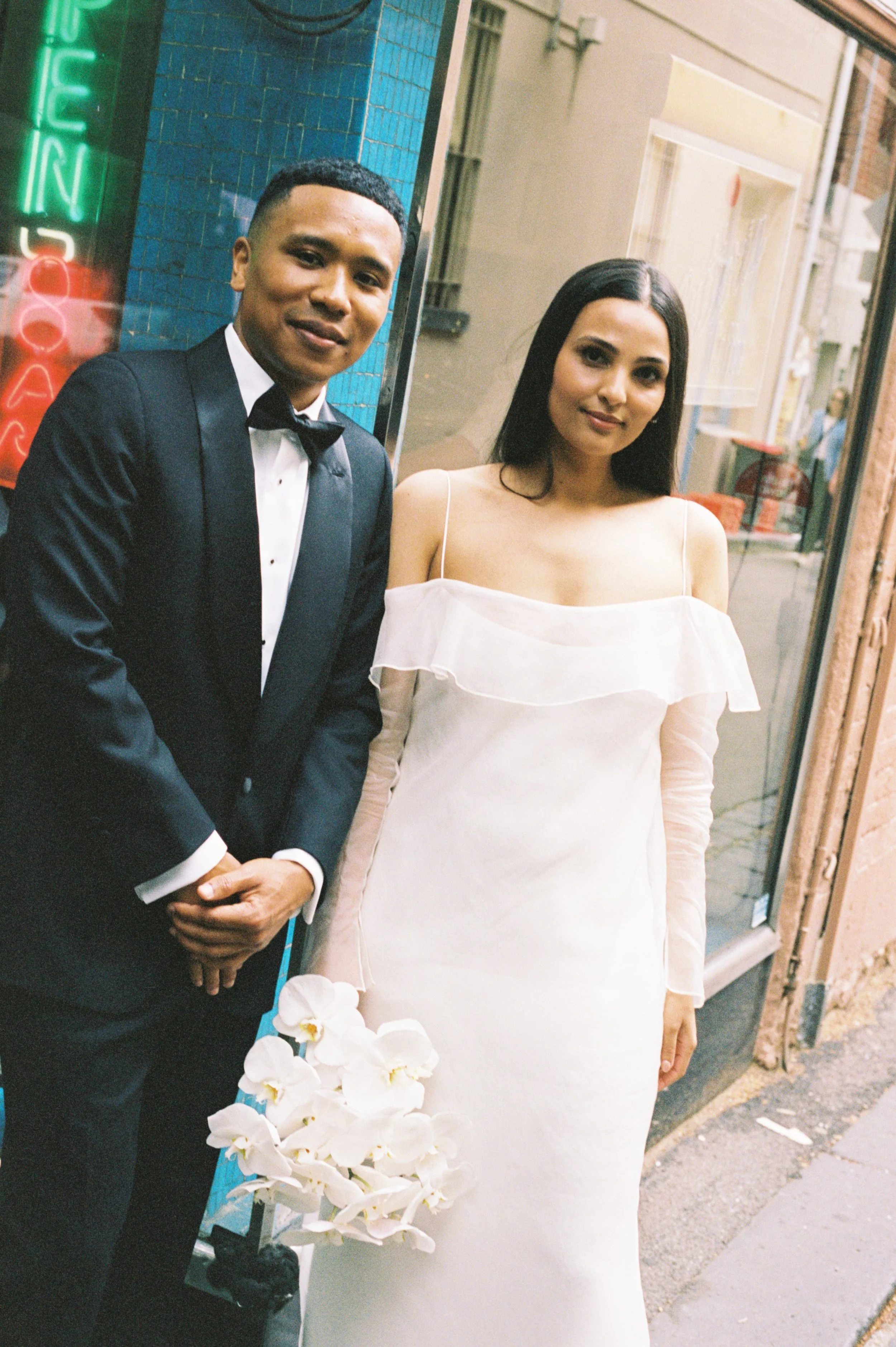 A newlywed couple standing outside of a shop, the groom in a black tuxedo with a bow tie, the bride in a white off-shoulder dress holding a bouquet of white flowers.