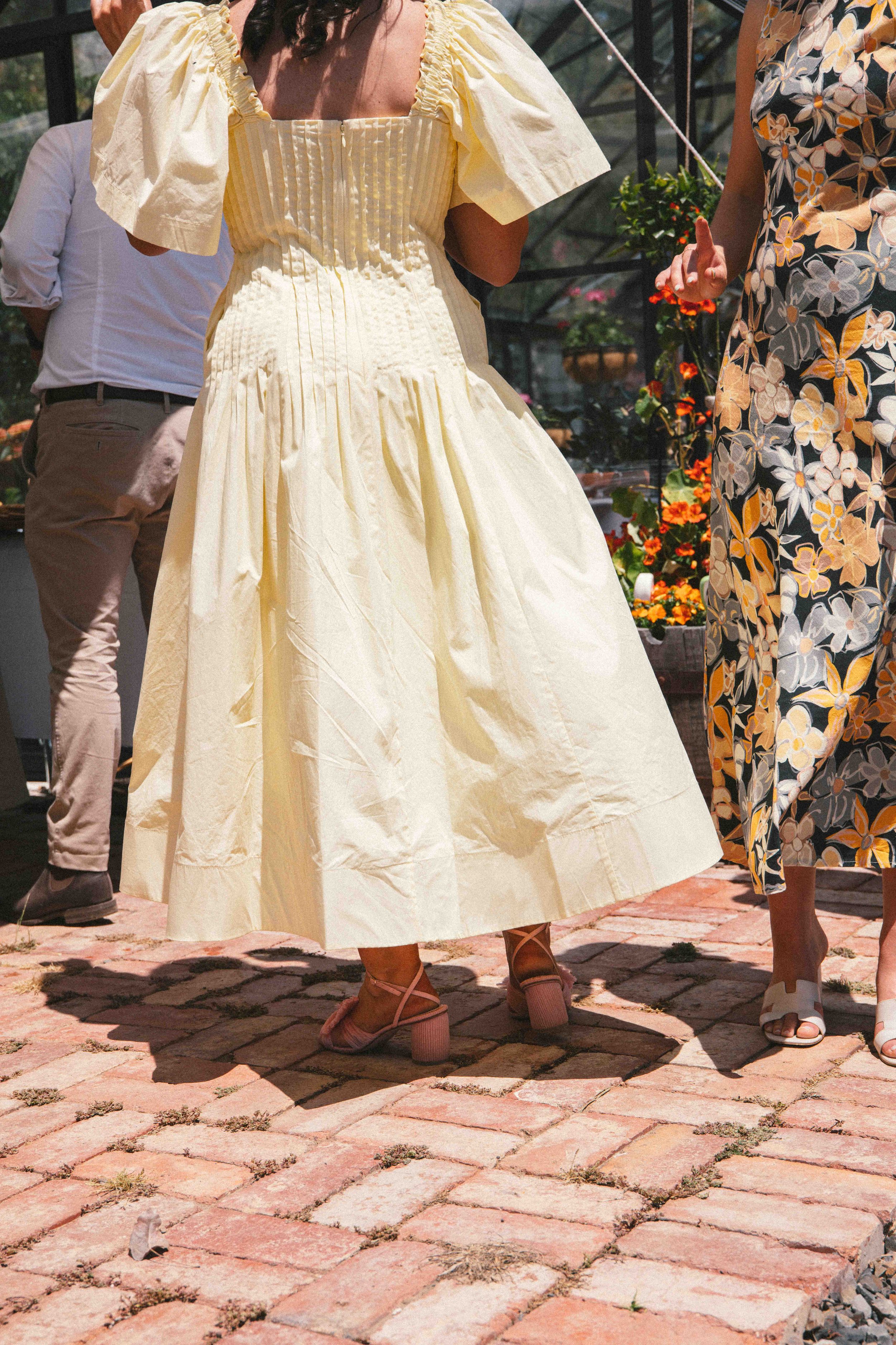 People standing outside on a brick patio, including a woman in a yellow dress with puffy sleeves and block-heeled shoes, at a garden or plant nursery.
