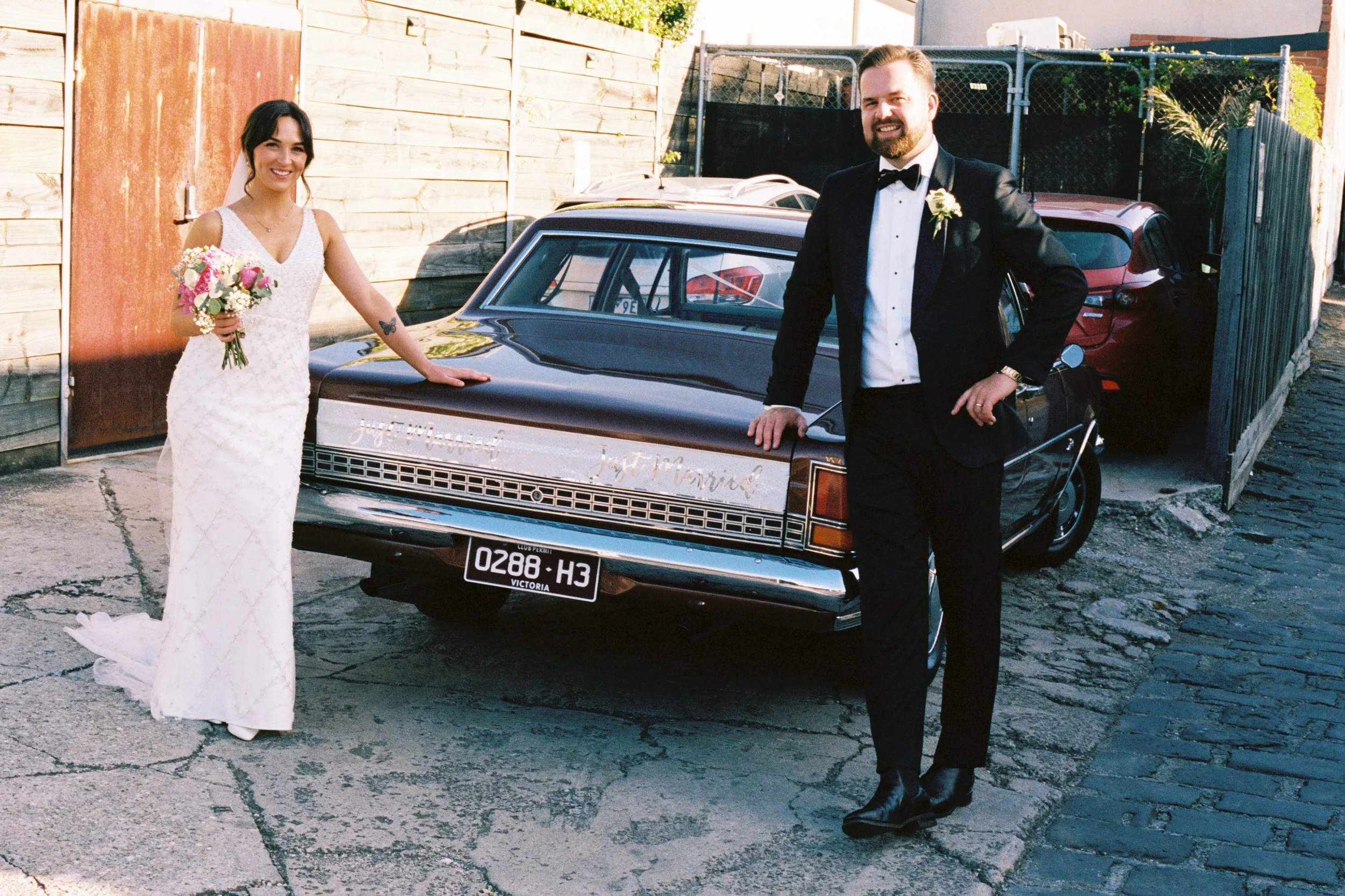 A bride and groom in wedding attire standing outside near a vintage black car with a decorative badge, a wooden fence, and parked cars in the background.