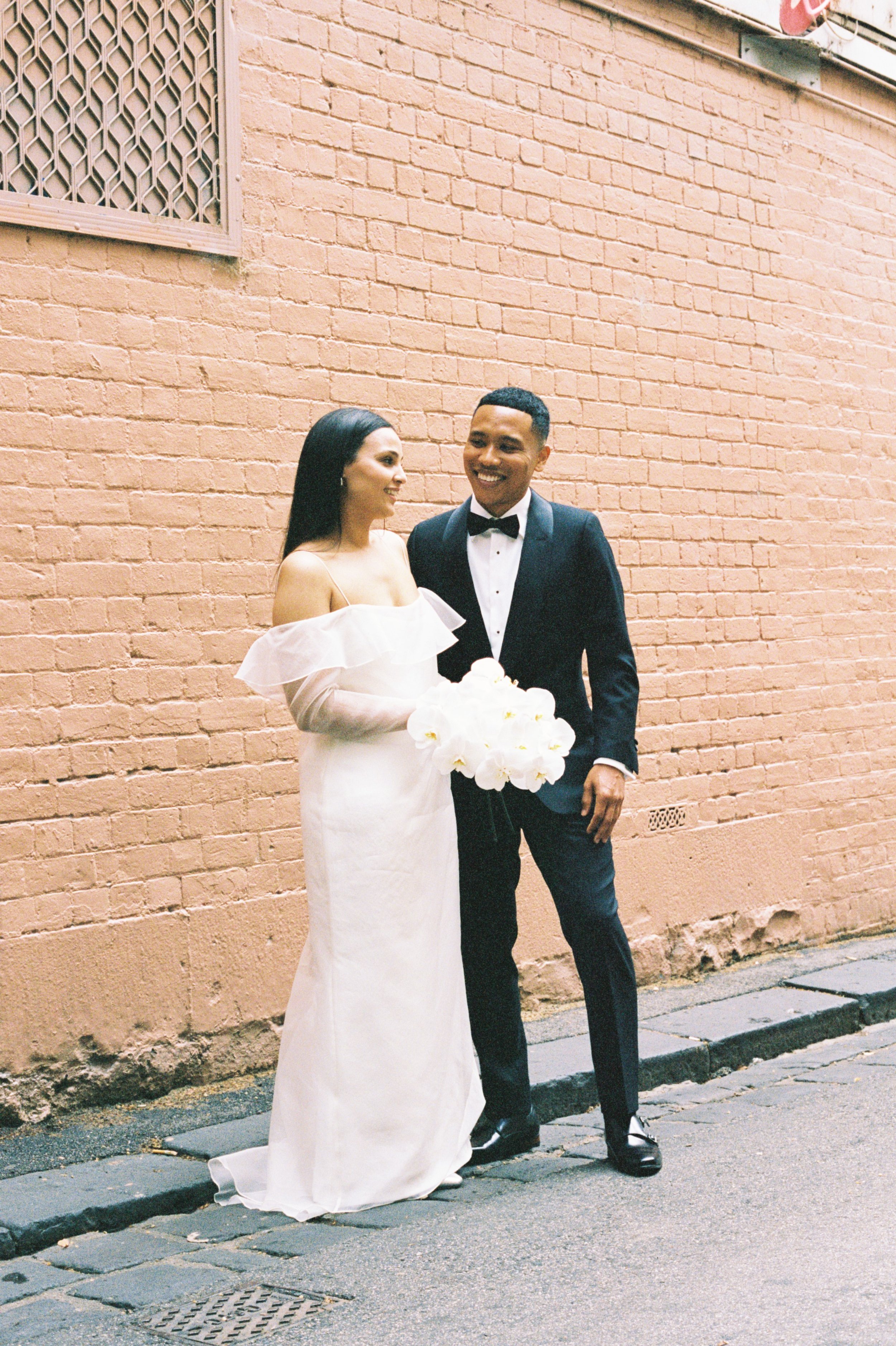 A bride and groom in wedding attire standing on a sidewalk against a pink brick wall, smiling and looking at each other. The bride is holding a bouquet of white flowers, wearing a white dress with off-shoulder ruffles, and the groom is in a black tux