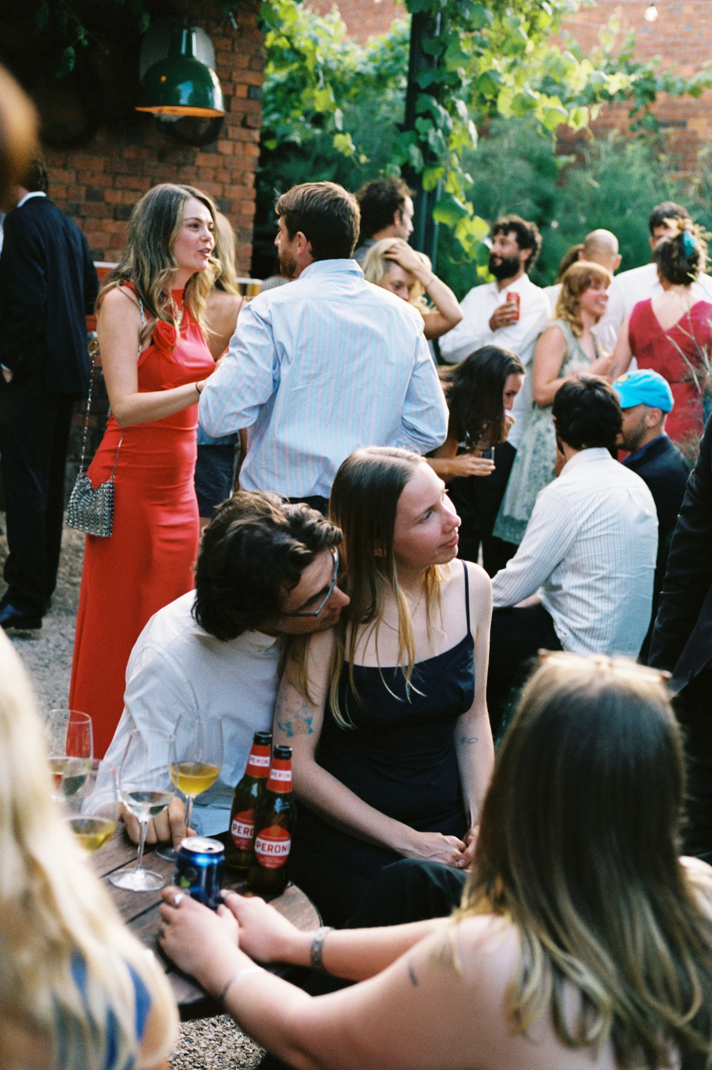 People socializing at an outdoor gathering, with some standing and talking, and others sitting at a table with drinks. The setting is lush and green, with a brick wall and vines in the background.