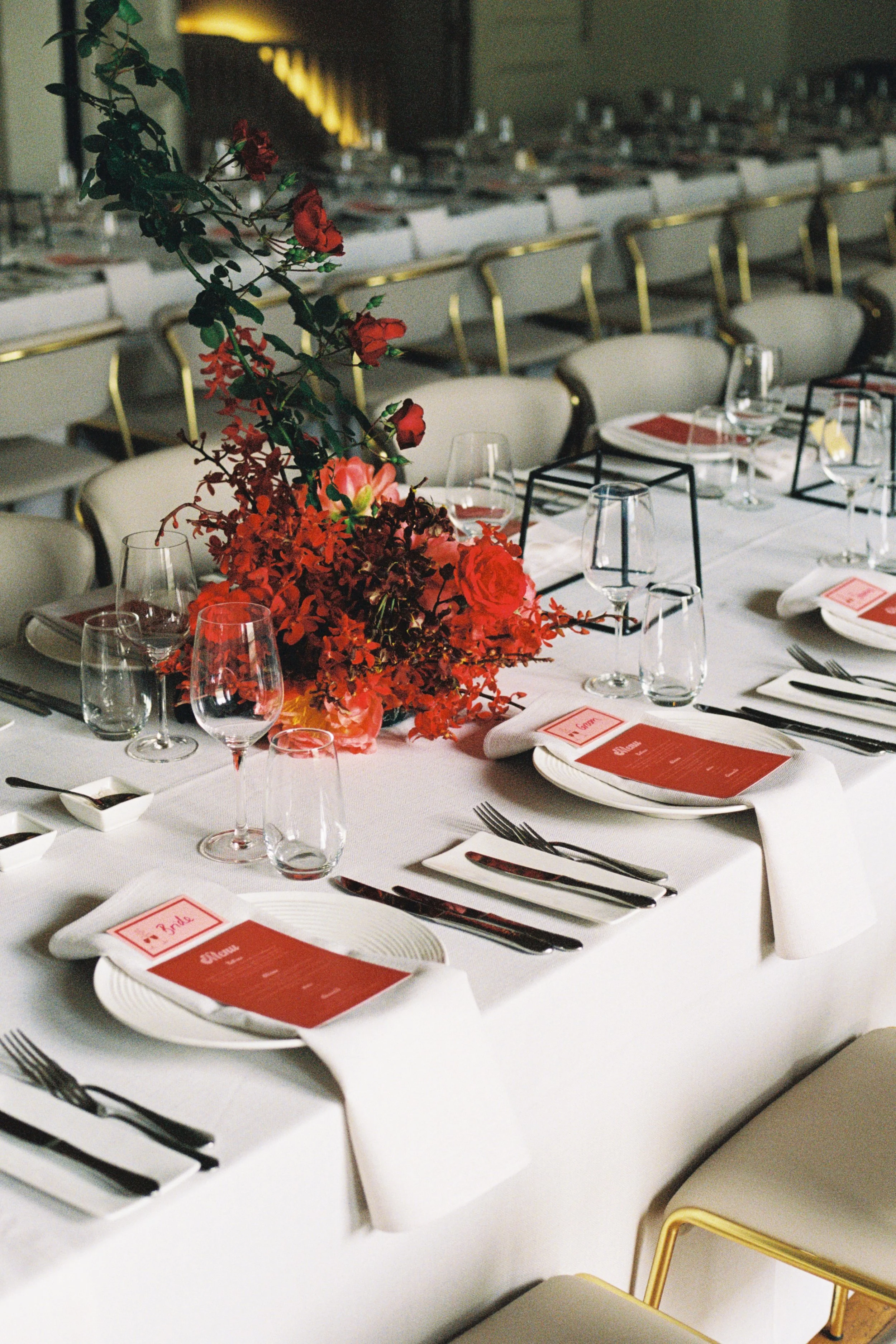 Table set for a formal event with a red floral centerpiece, white tablecloth, plates, silverware, glassware, and red menus, in a dimly lit banquet hall.