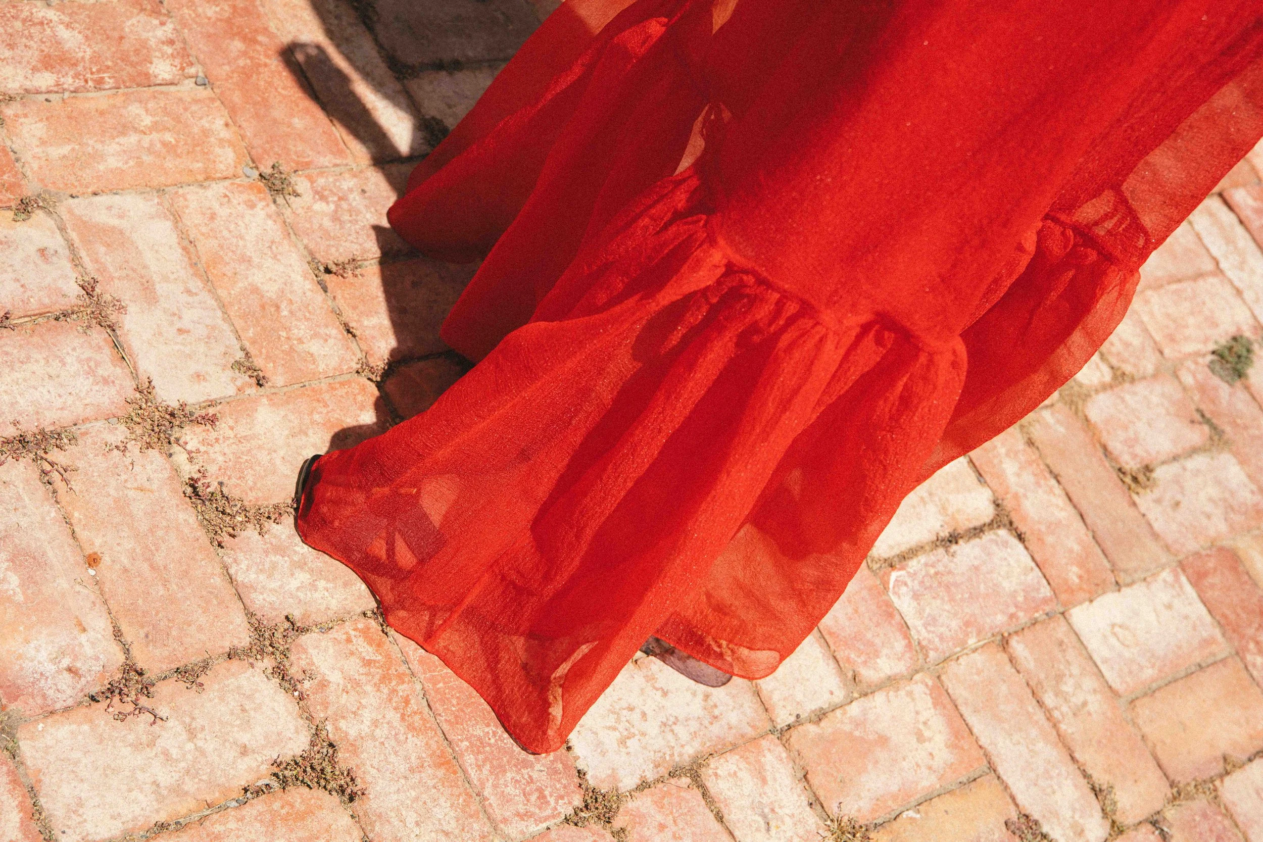 The bottom part of a person wearing a long, sheer red dress standing on a brick sidewalk, with sunlight casting shadows.