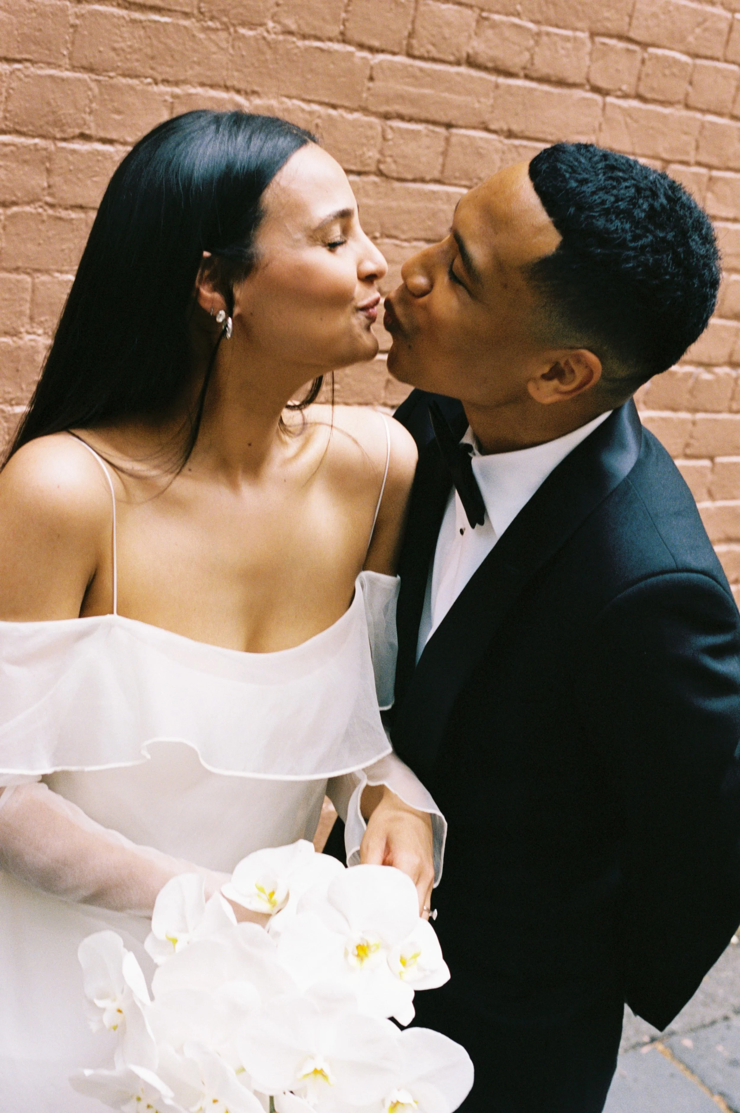 A bride and groom sharing a kiss, with the bride holding a bouquet of white orchids, standing against a brick wall.