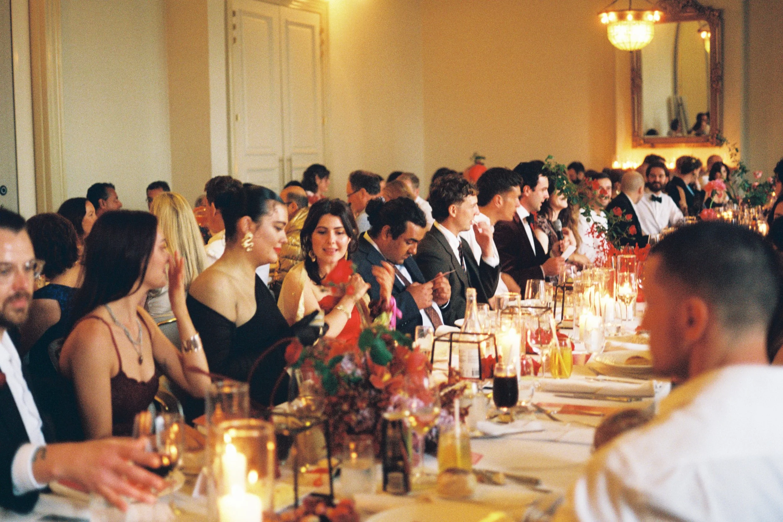 People dressed in formal attire seated at a long banquet table during a wedding reception or formal event, with candles and floral centerpieces.