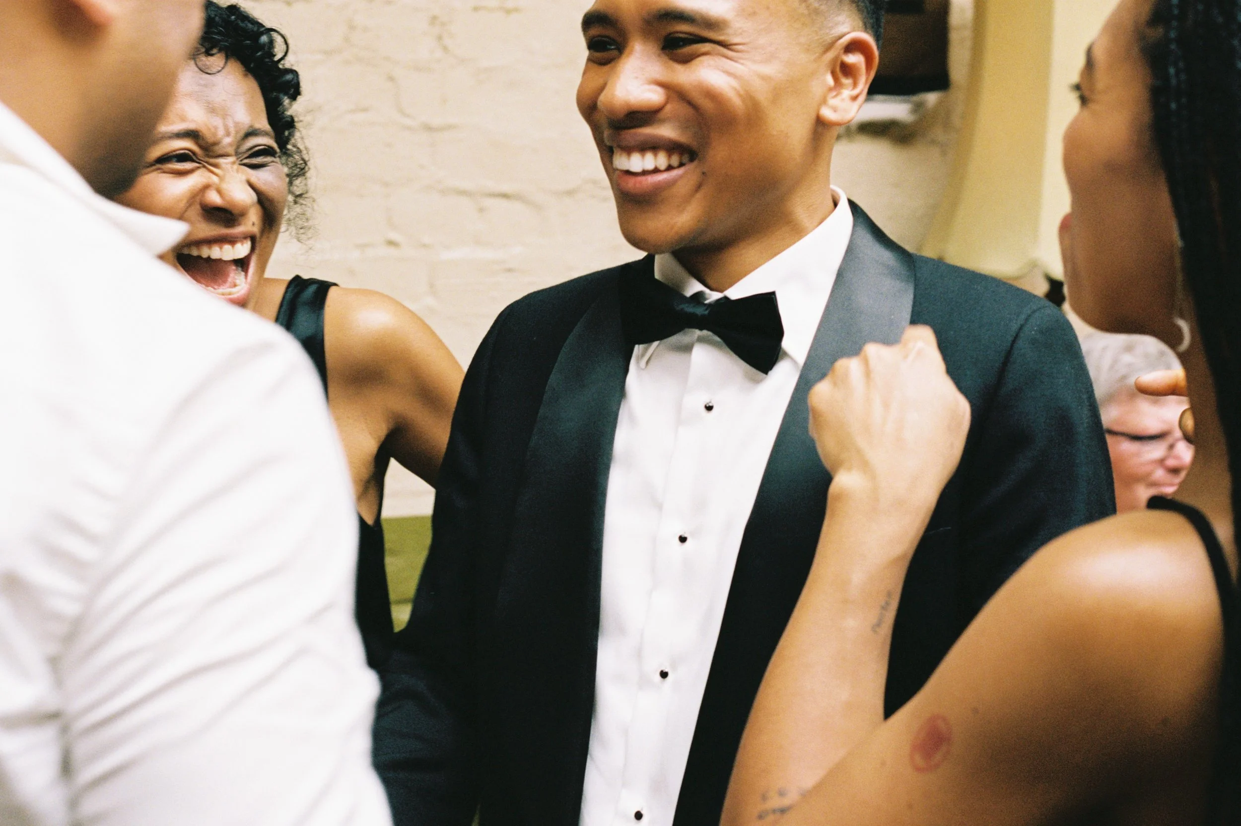 Smiling man in tuxedo and bow tie surrounded by women at a celebration or party. Wedding photography Melbourne film analogue candid.