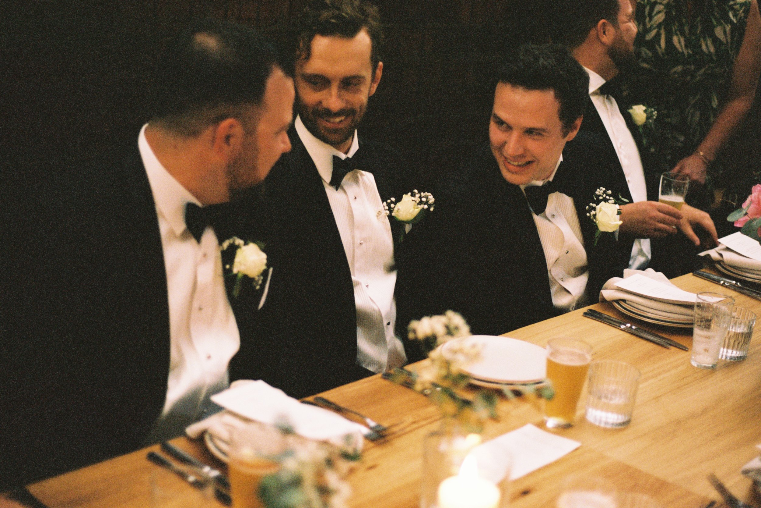 Four men in tuxedos sitting at a wedding reception table, smiling and talking, with table settings and drinks visible.