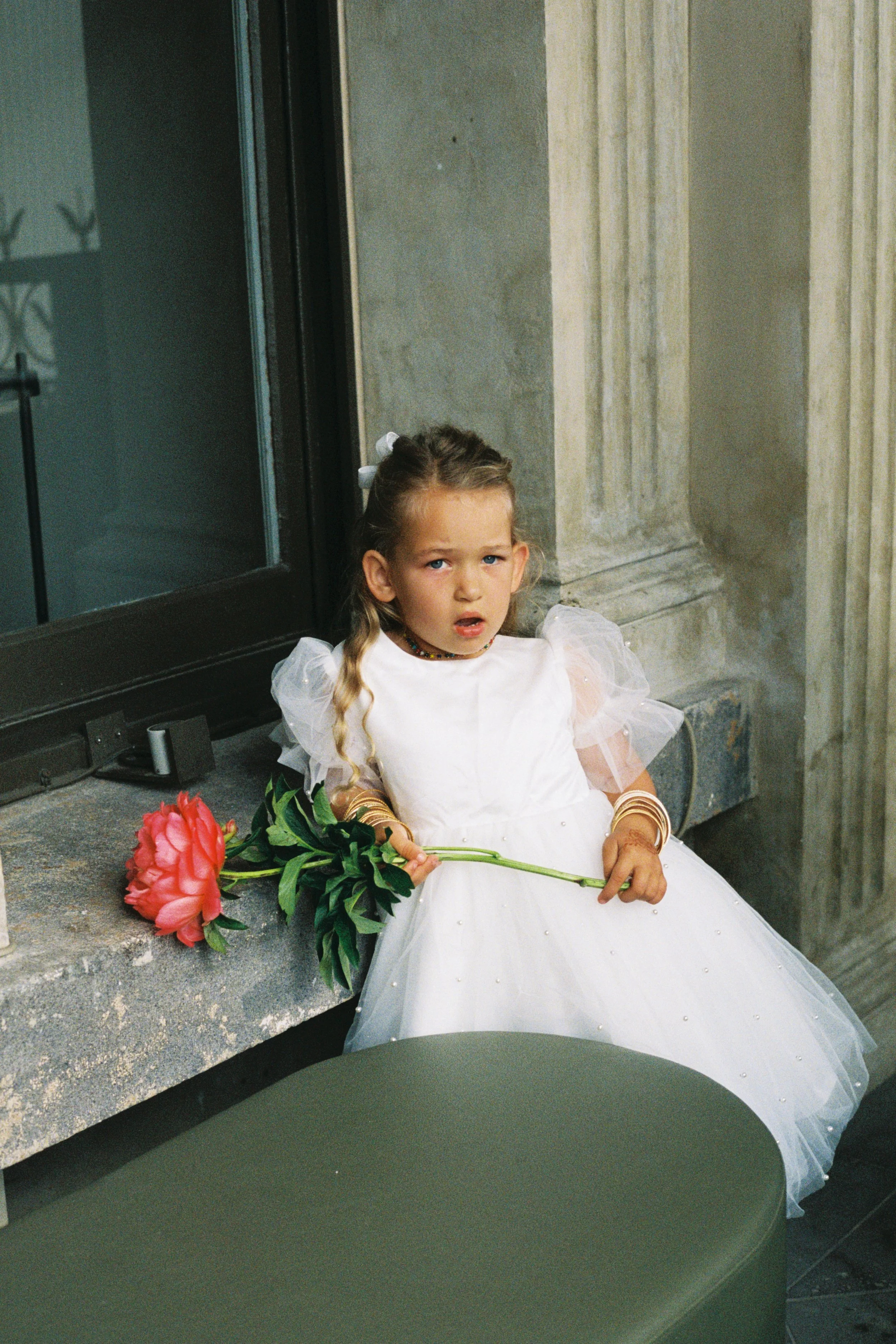 Young girl in a white dress sitting on a stone ledge holding a pink flower, with a surprised or confused expression, indoors near a window.