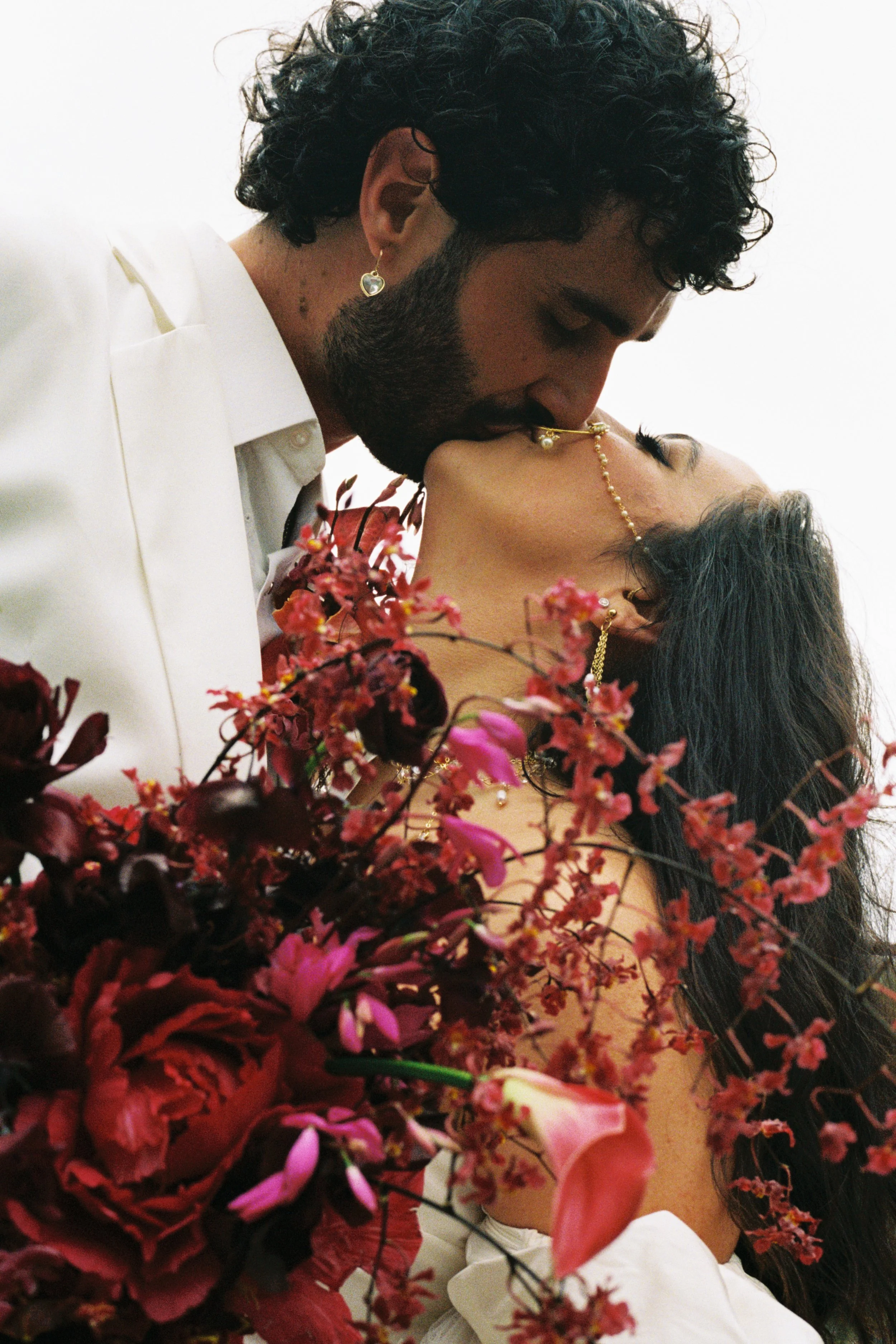 A man with dark curly hair and a beard is kissing a woman who has long dark hair; she is surrounded by pink and red flowers and is wearing gold jewelry, including earrings, a nose chain, and a necklace.