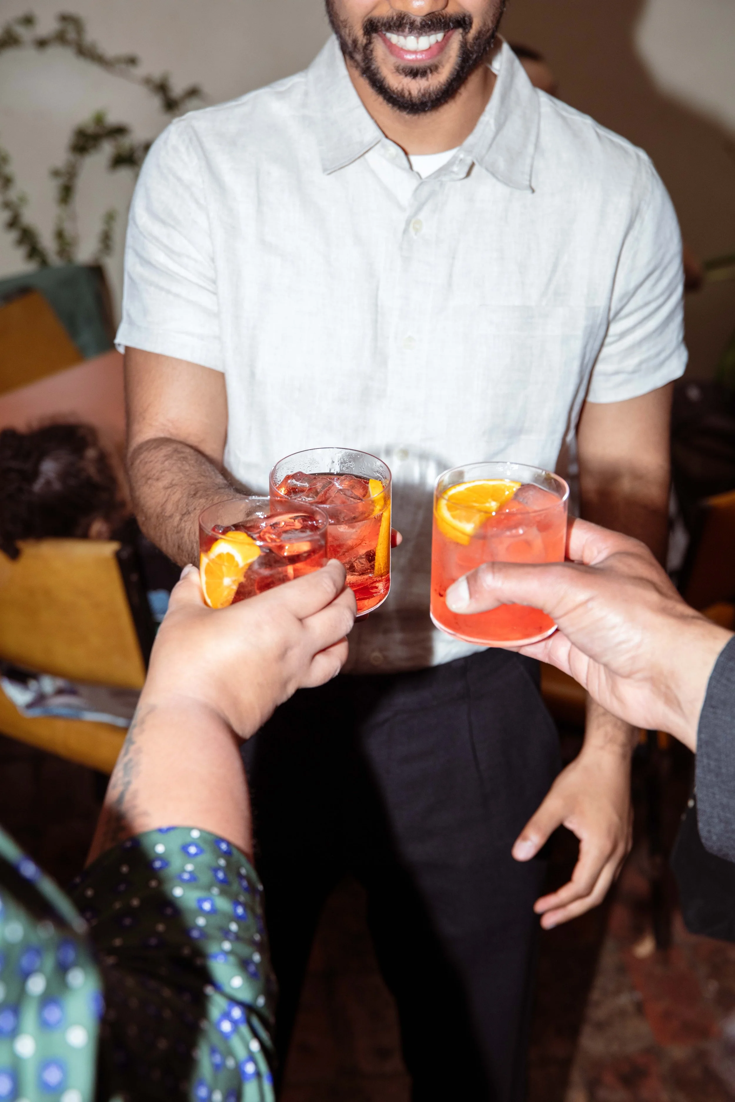 Three people clinking glasses of pink cocktails with orange slices, with a smiling man in a white shirt in the background.