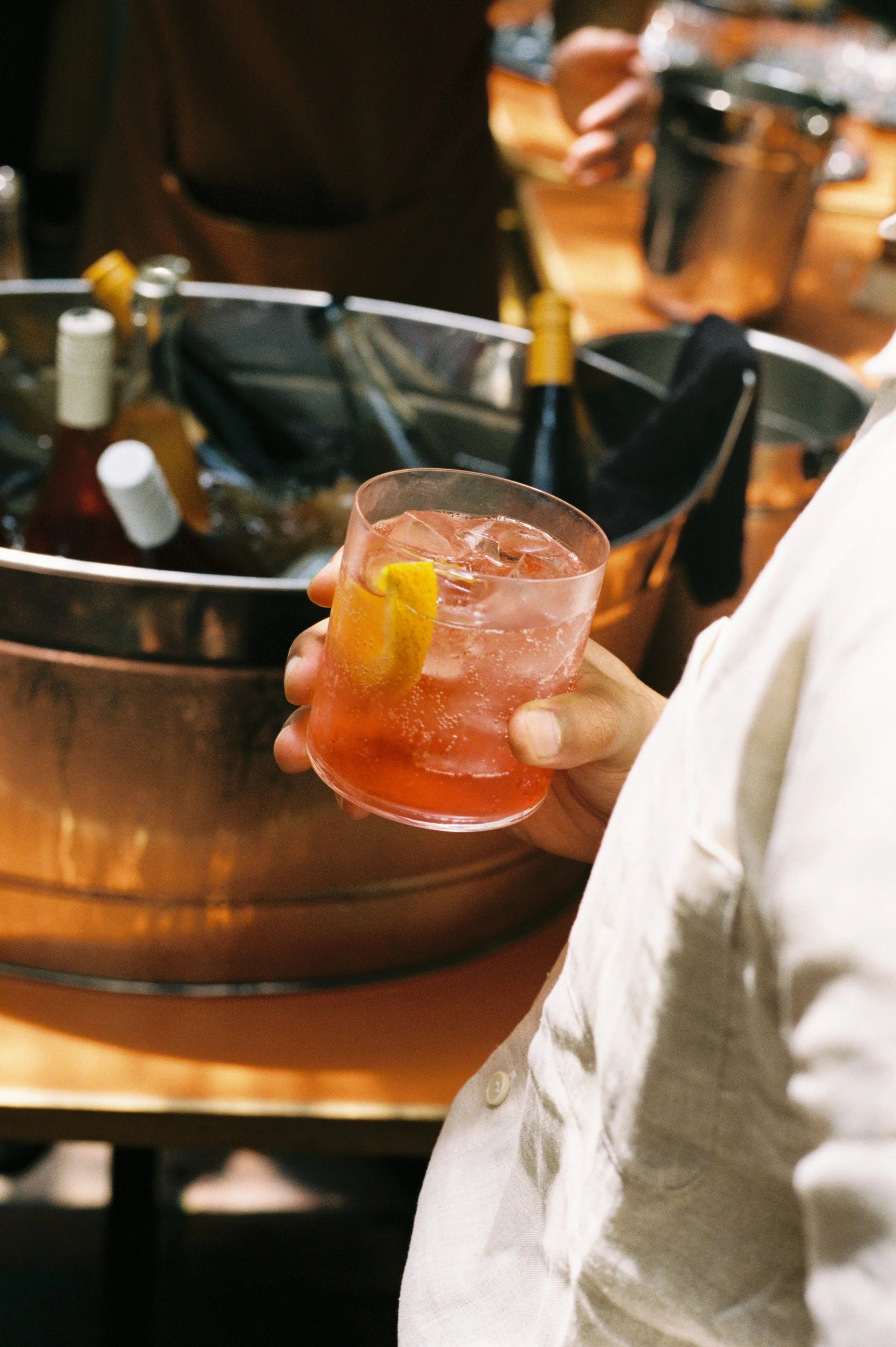 Person holding a glass of pink cocktail with a lemon wedge in a bar or restaurant setting.