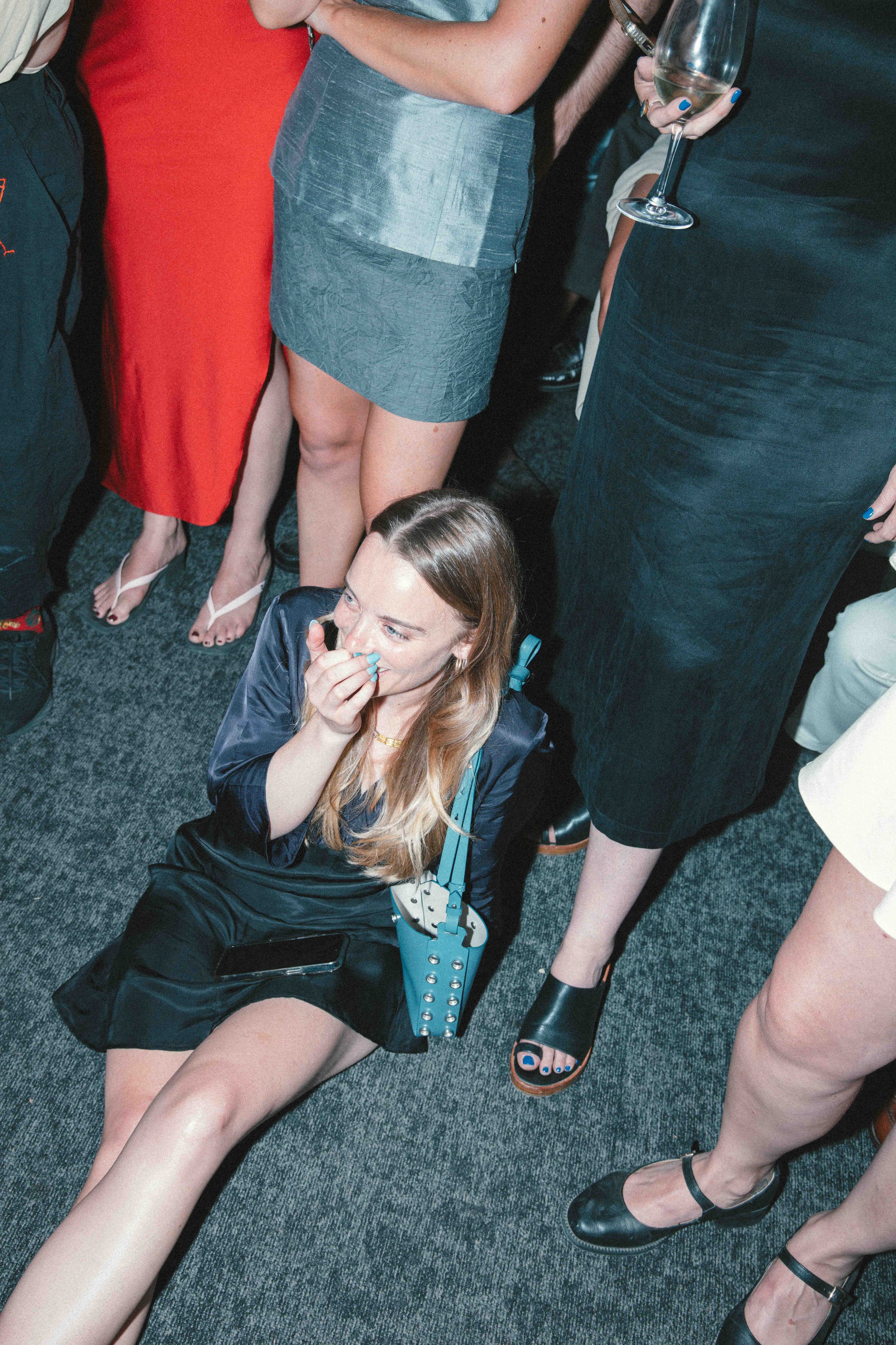A woman sitting on the floor at a social gathering, laughing and covering her mouth with her hand. She is surrounded by standing individuals wearing dresses and heels, with some holding drinks.