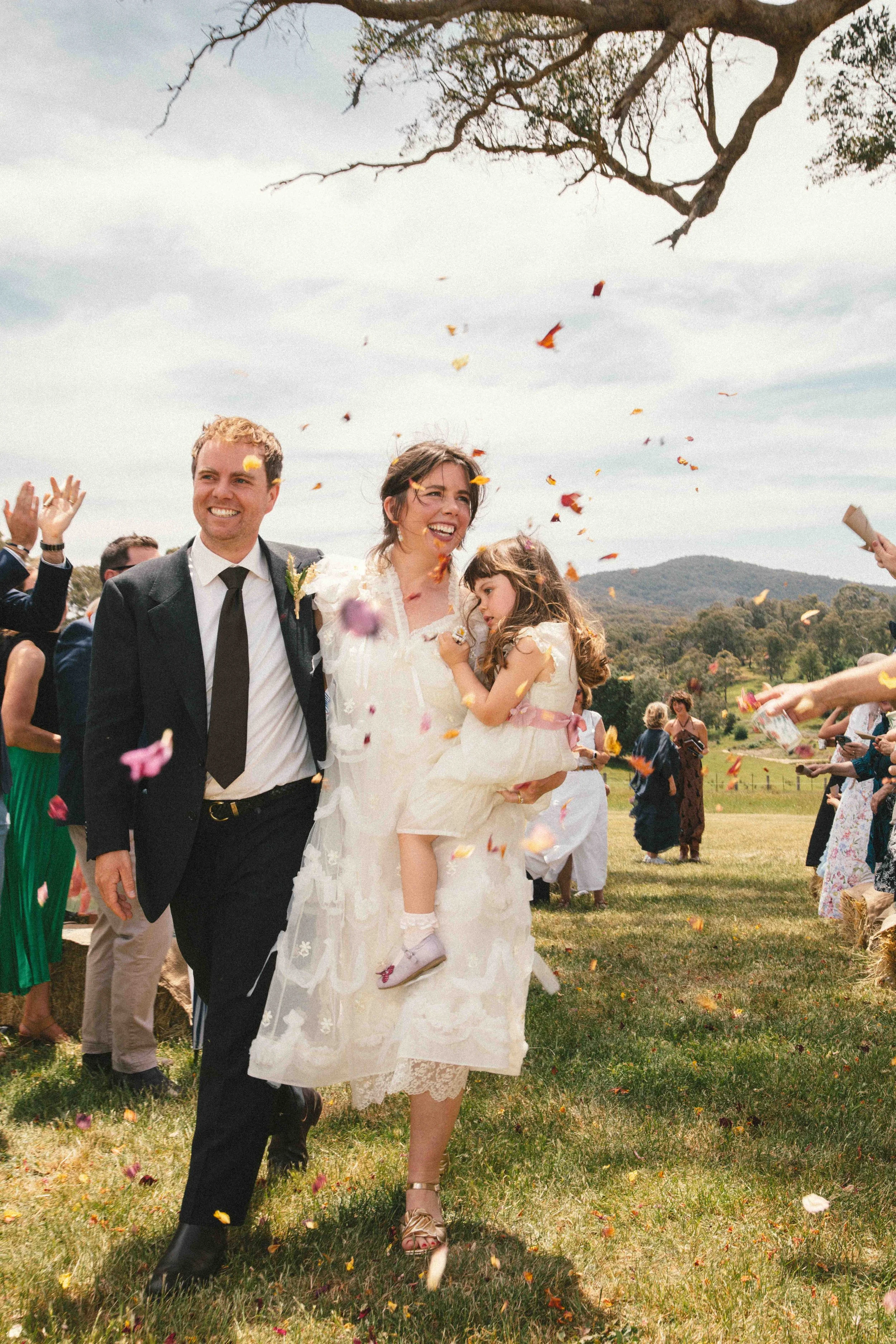 A newlywed couple walking outdoors on their wedding day, surrounded by guests throwing flower petals. The groom is in a black suit and the bride in a white wedding dress holding a young girl in white dress and pink sash. The background shows a grassy