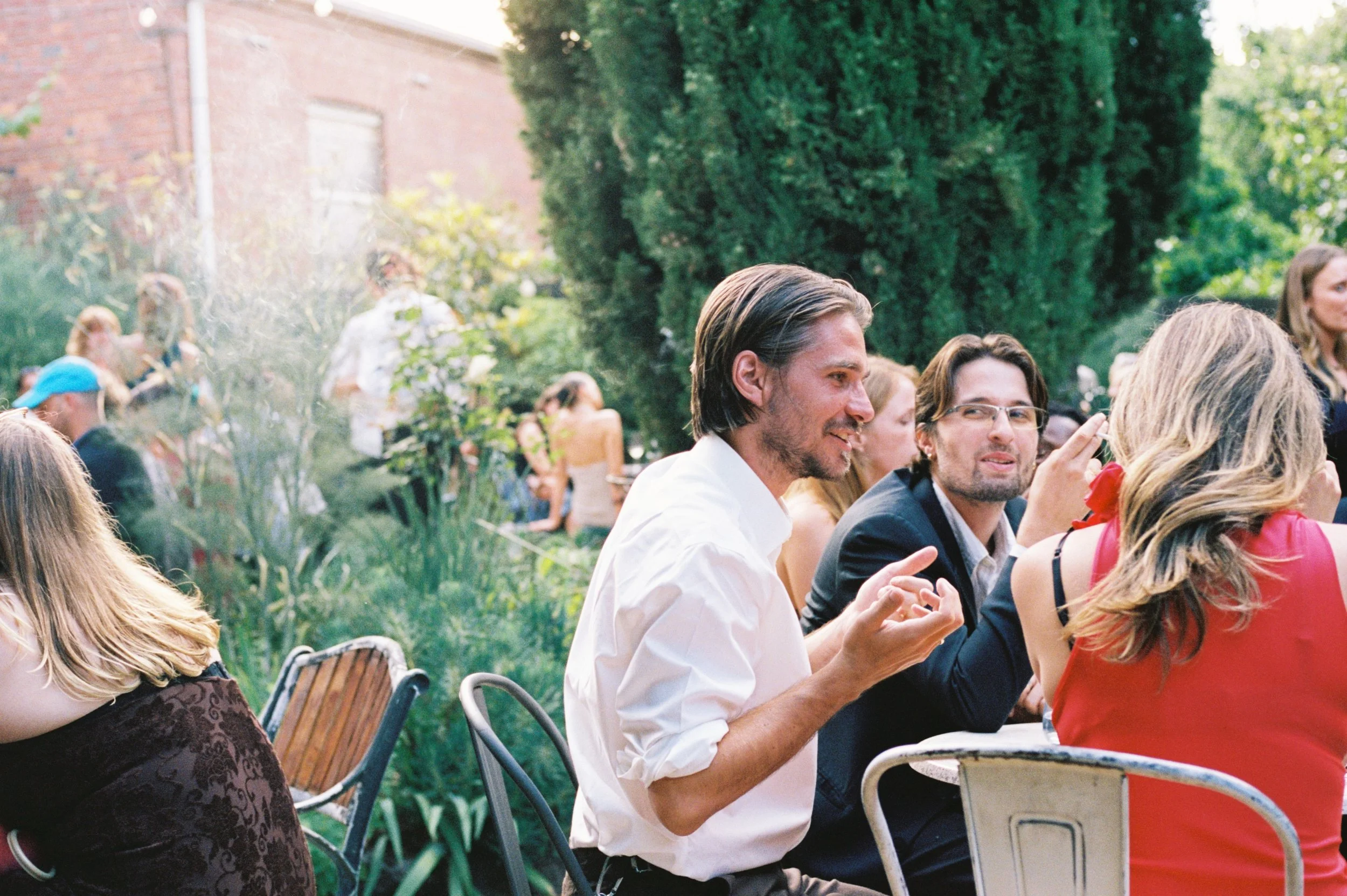 People gathered at an outdoor garden party, sitting on chairs and enjoying conversations amidst greenery and sunlight.