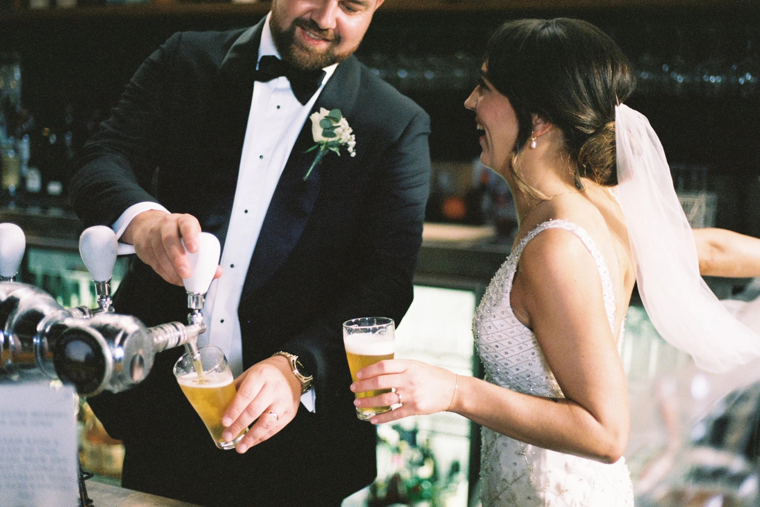 A newlywed couple at a bar, with the groom pouring beer into a glass while the bride holds her own glass, both smiling and dressed in wedding attire.