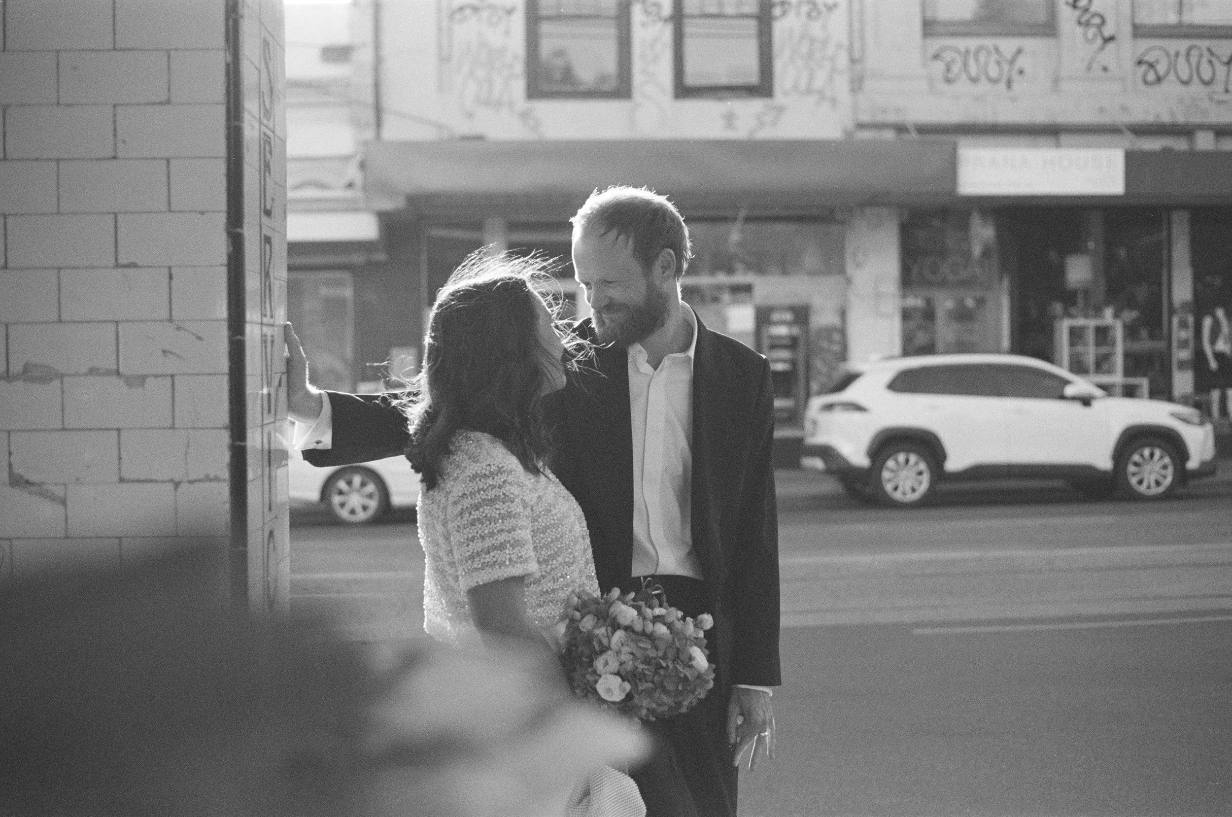 A black and white photo of a couple standing on a city sidewalk, smiling at each other, with the woman holding a bouquet of flowers. The man has his arm resting on a wall beside them, and a car and storefronts are visible in the background.