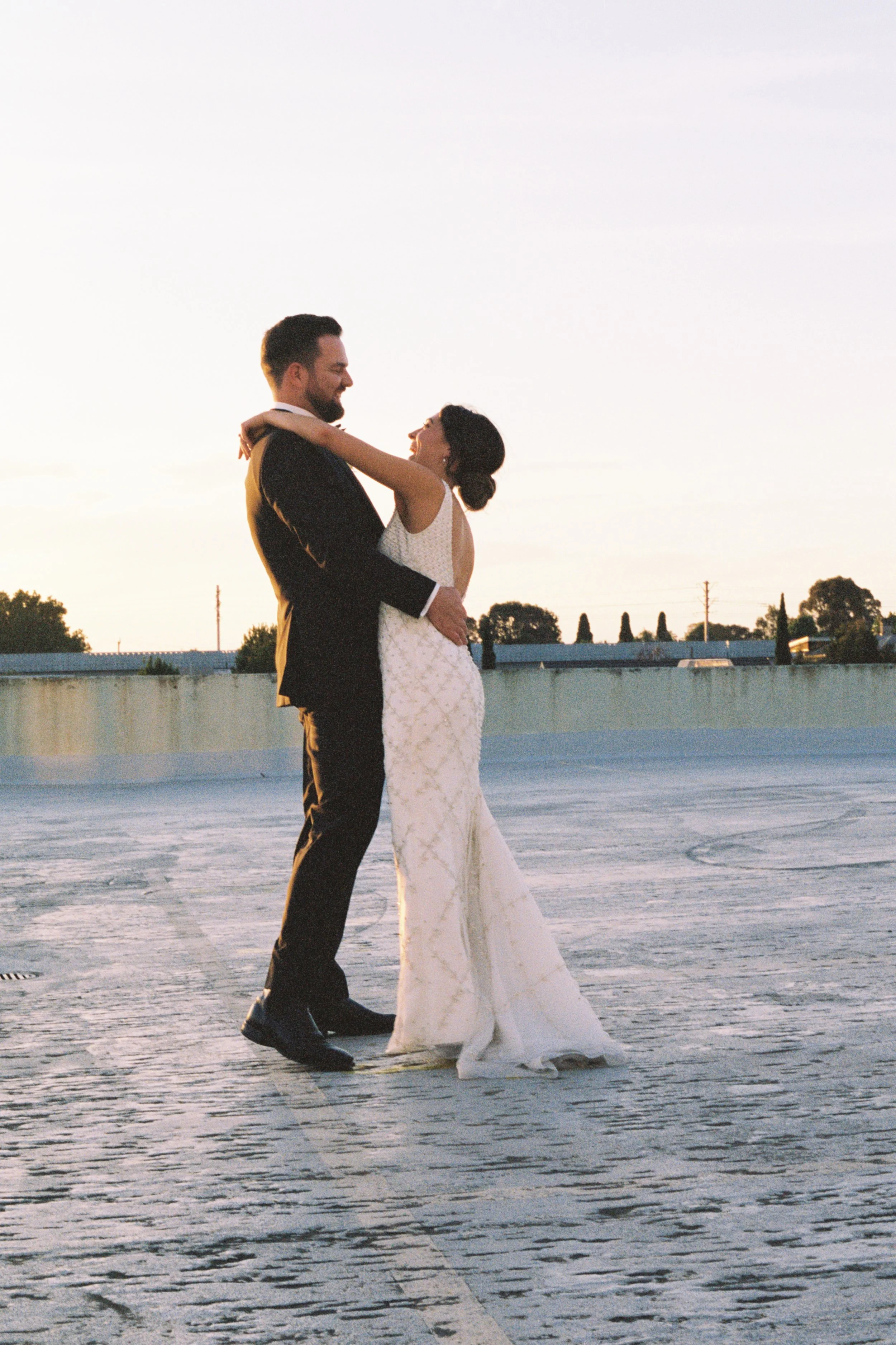 A bride and groom embrace while dancing outside during sunset, facing each other with smiles