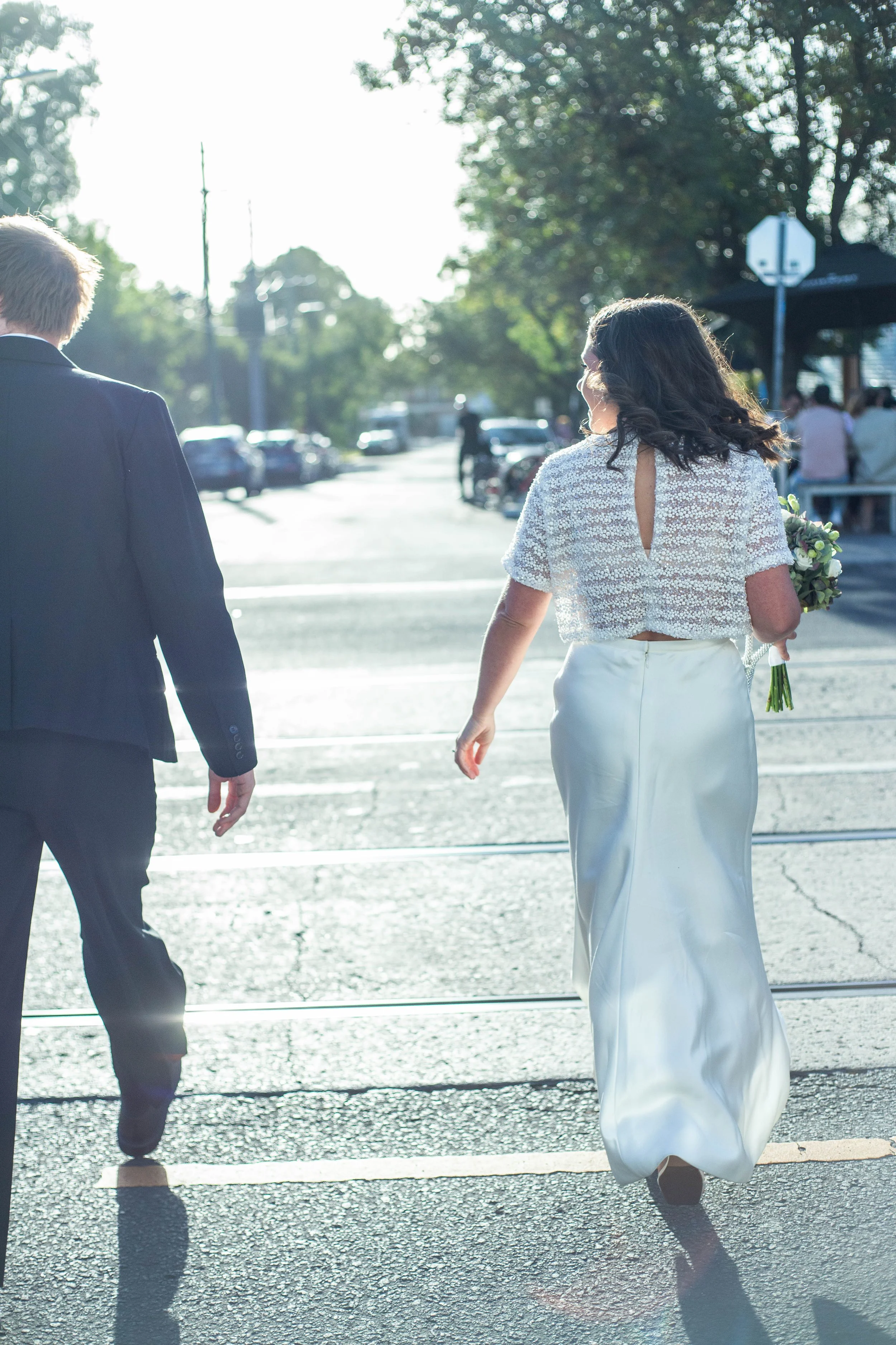 A woman in a white wedding dress holding a bouquet walking across a crosswalk beside a man in a suit, backlit by the sun on a city street.