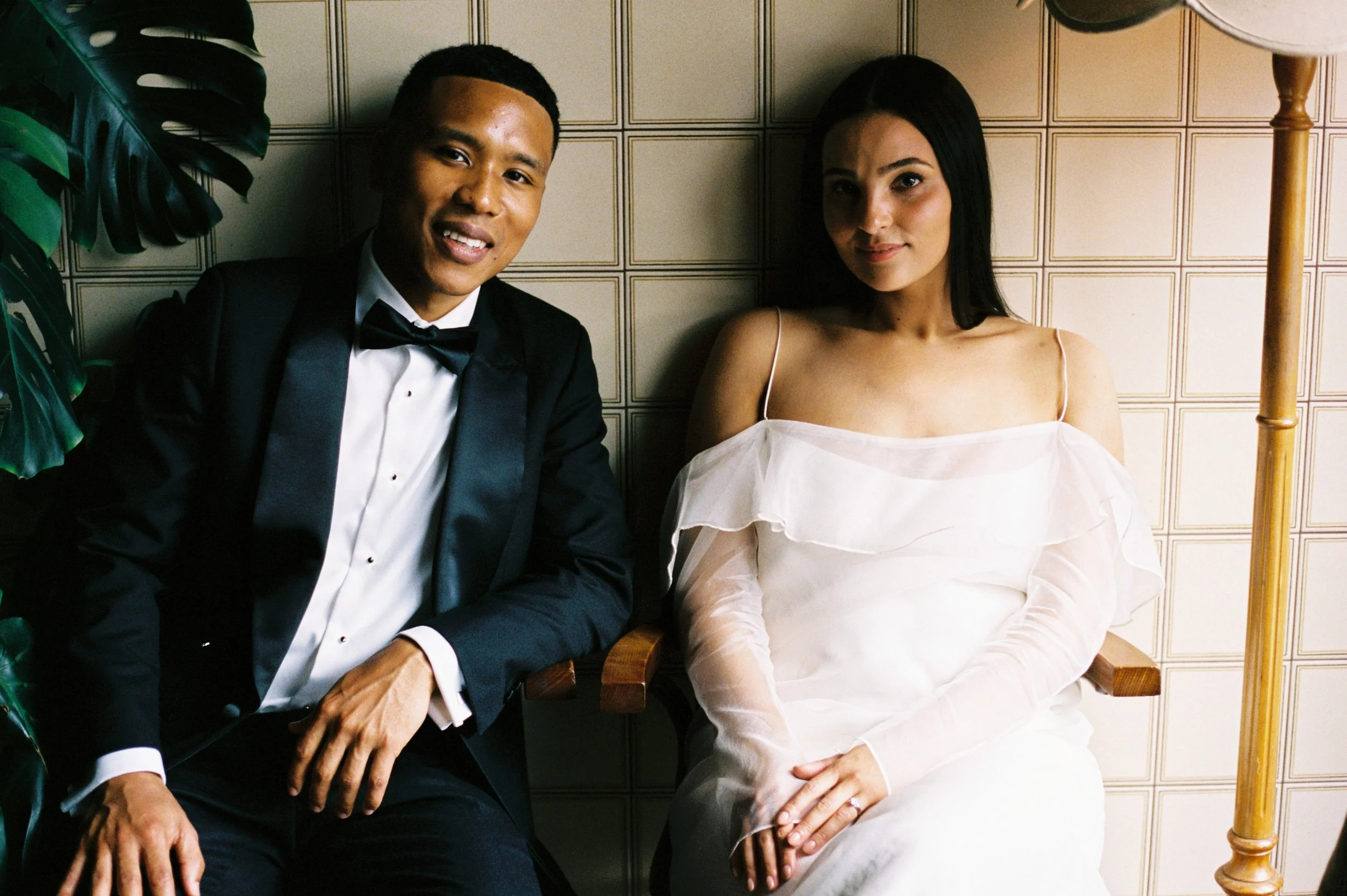 A man in a tuxedo sitting next to a woman in a white off-shoulder dress, both sitting against a tiled wall, with a lamp and a leafy plant nearby.