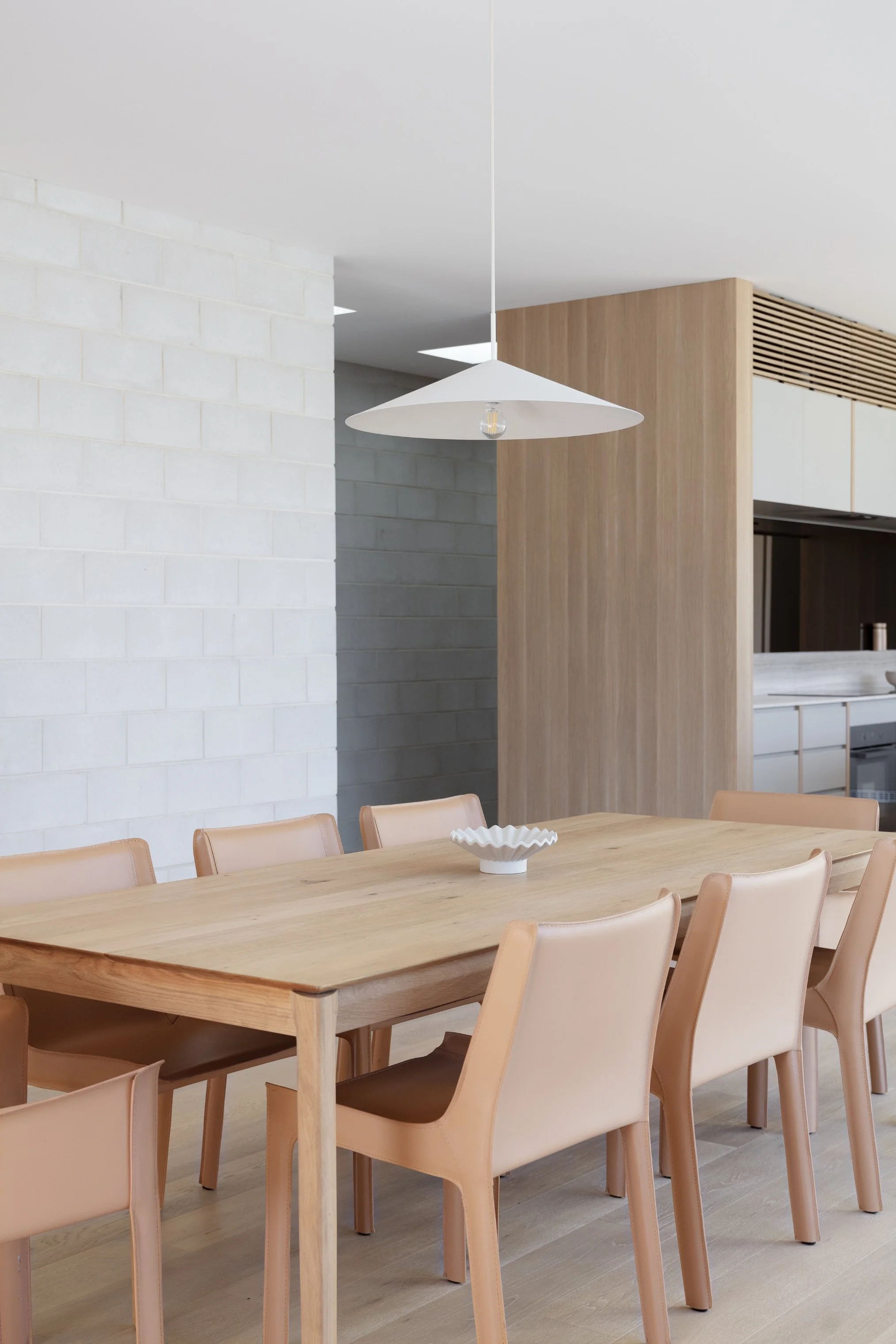 A minimalist dining room with a wooden table, beige chairs, a white pendant lamp hanging from the ceiling, and a modern kitchen in the background with light-colored cabinets and a wooden accent wall.