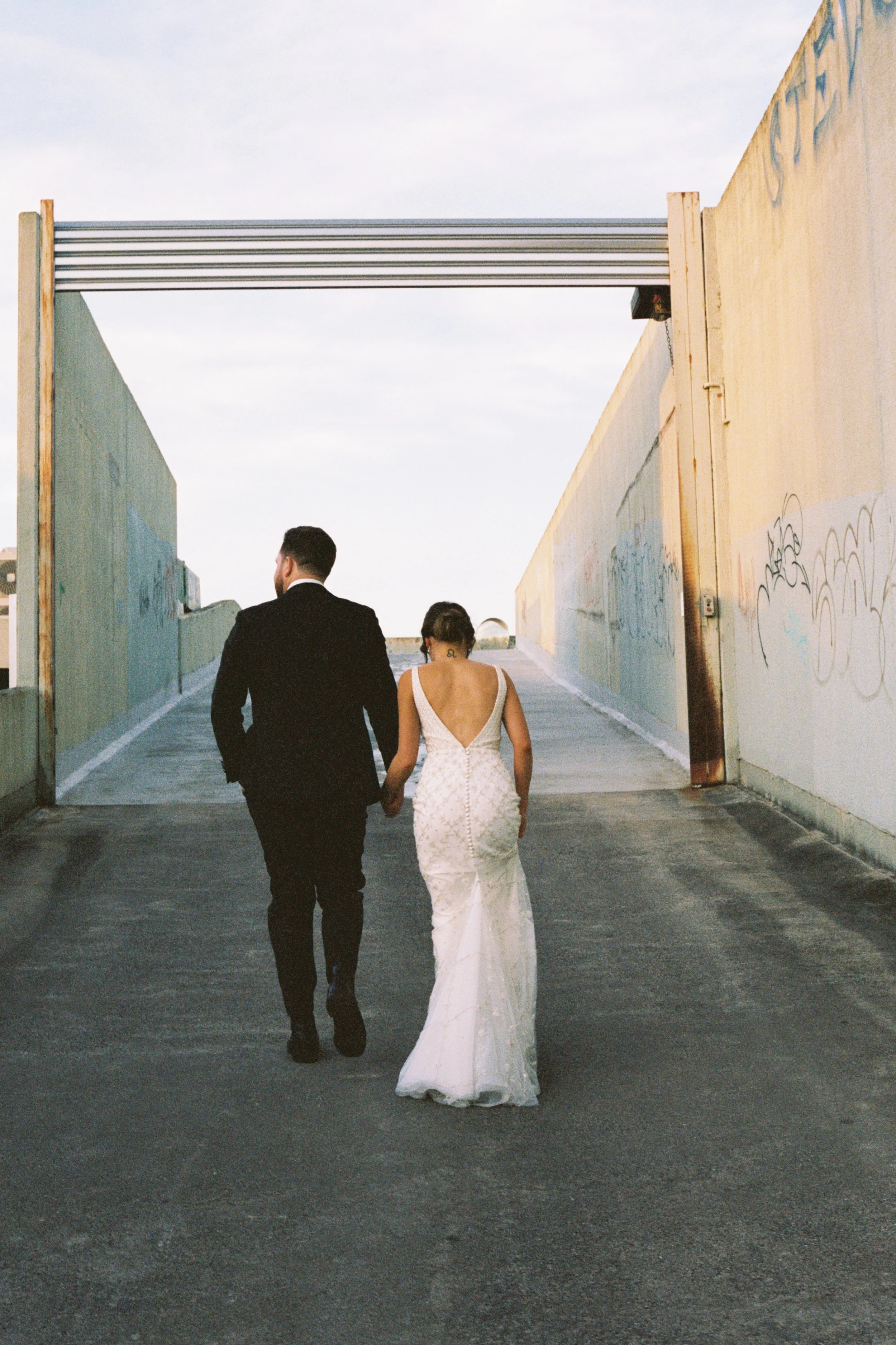 A newlywed couple, dressed in formal attire, holding hands and walking away, with a mural-covered wall on one side and an open sky overhead.