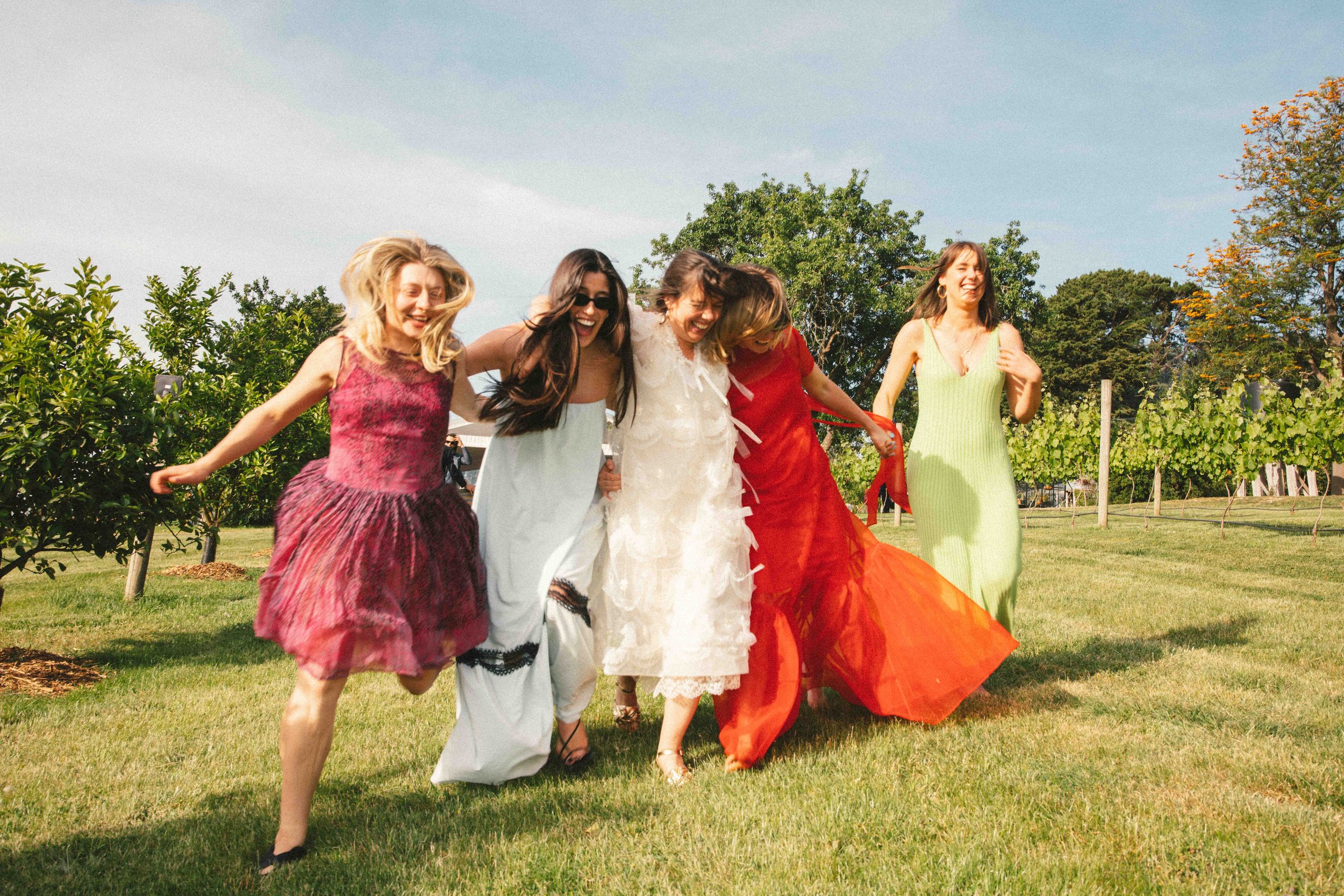 Group of five women in colorful dresses happily walking and laughing together on a sunny day in a grassy outdoor setting with trees in the background.