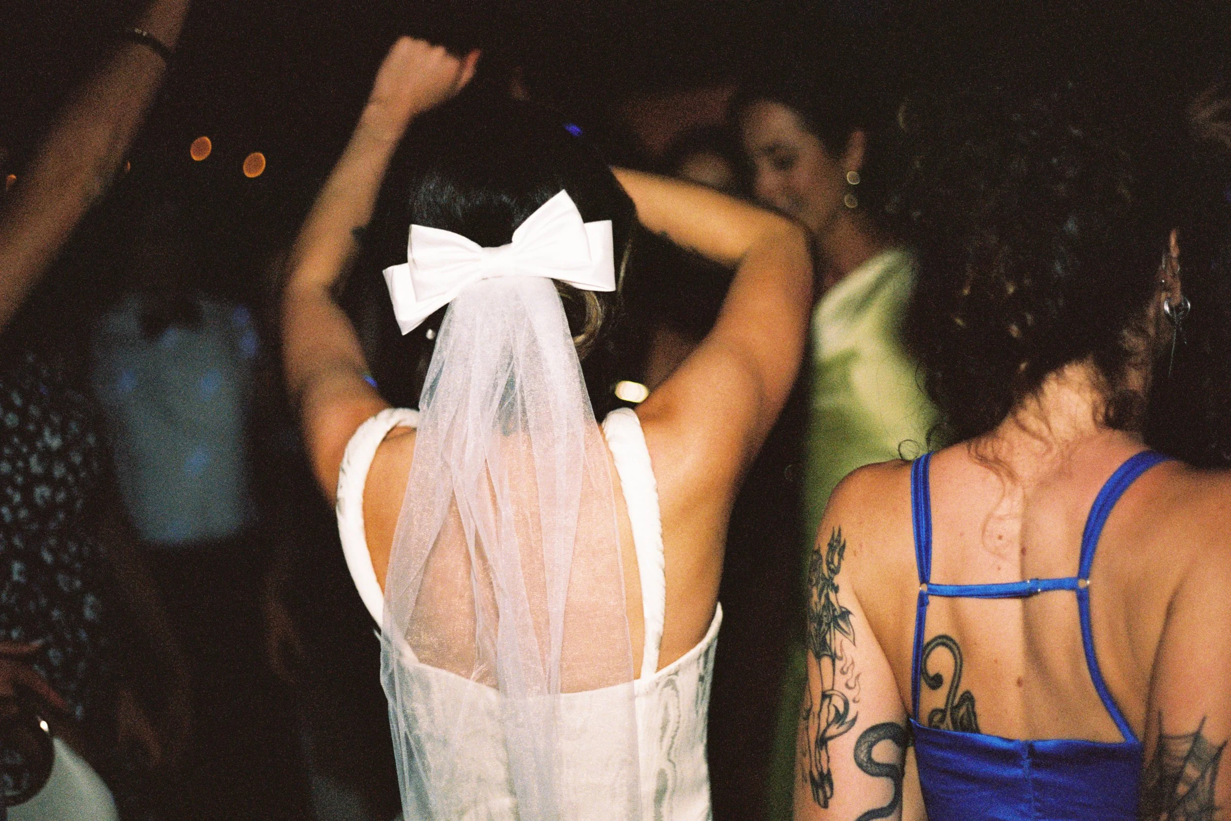 People dancing at a party, with the focus on a woman wearing a white veil with a large bow, suggesting a wedding celebration.