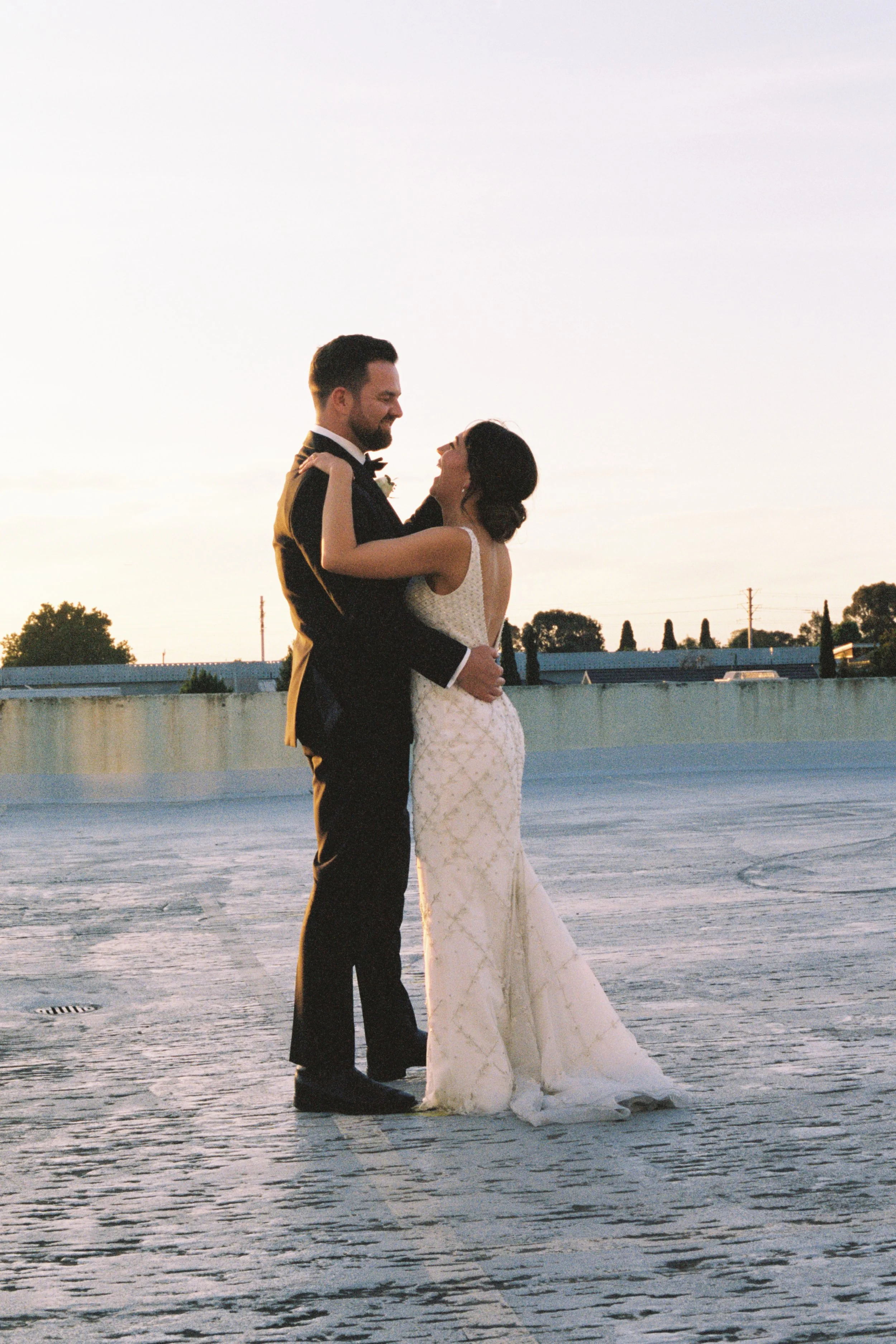 A bride and groom dancing on a rooftop at sunset, embracing each other and smiling.