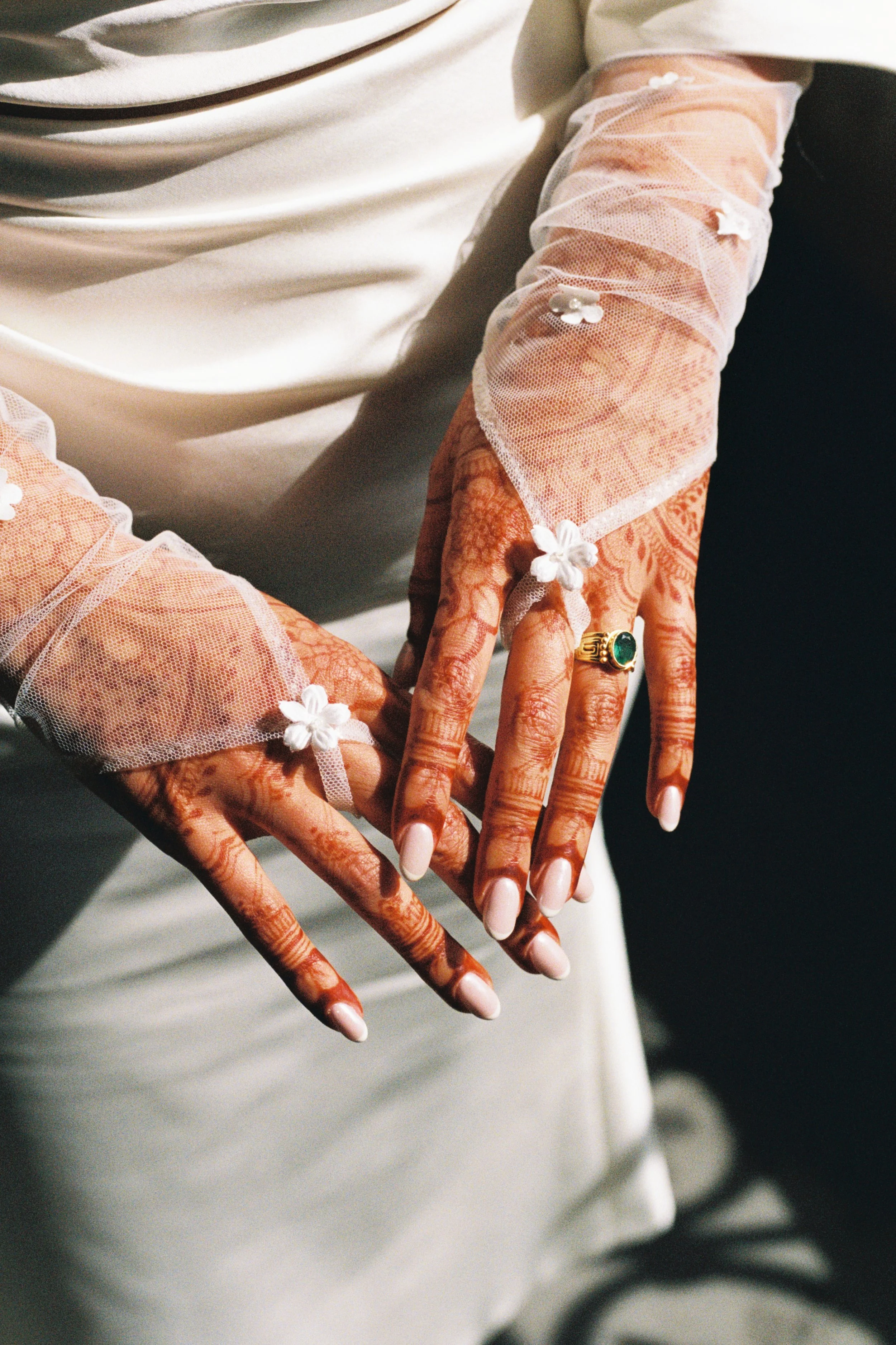 Close-up of hands with henna and mehndi designs, wearing a ring with green gemstone, and decorated with white floral accessories on sheer, white sleeves, against a dark background.