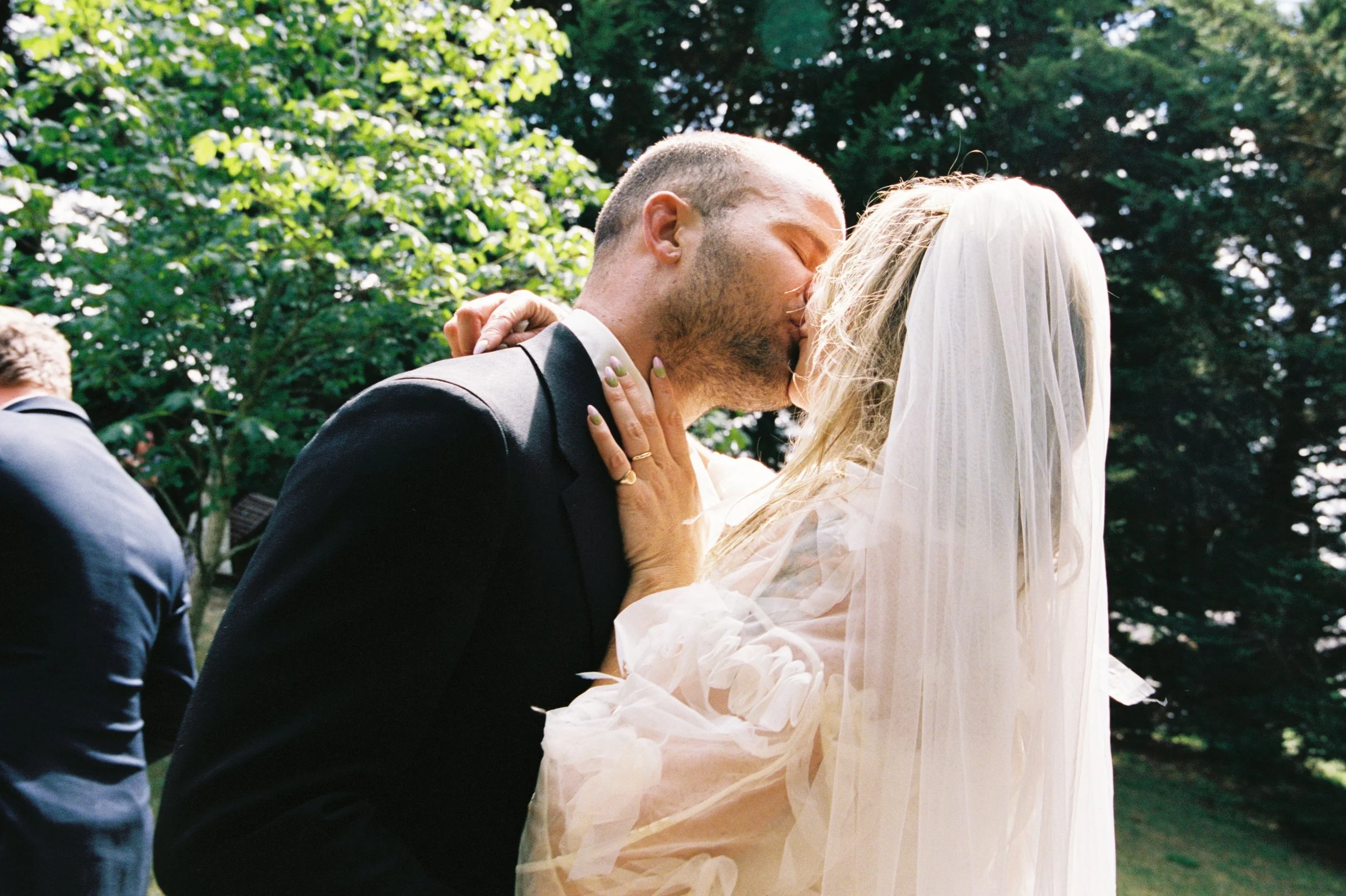 A couple kissing at their wedding outdoors, with the bride wearing a white veil and dress and the groom in a black suit, surrounded by green trees.