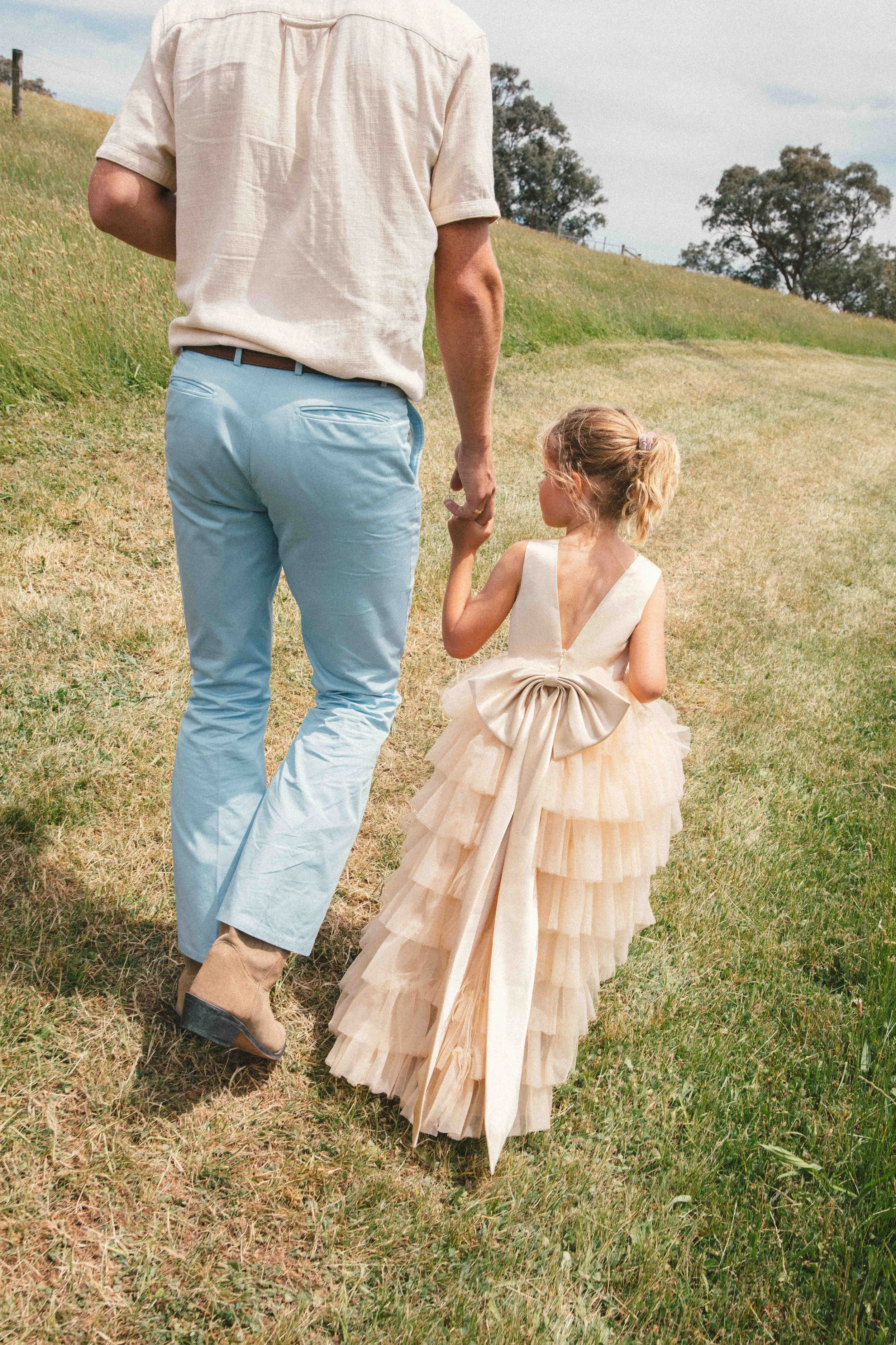 A man and a young girl walk hand in hand through a grassy field. The girl is wearing a tiered, cream-colored dress with a large bow on the back. The man is wearing light blue pants and a beige shirt.