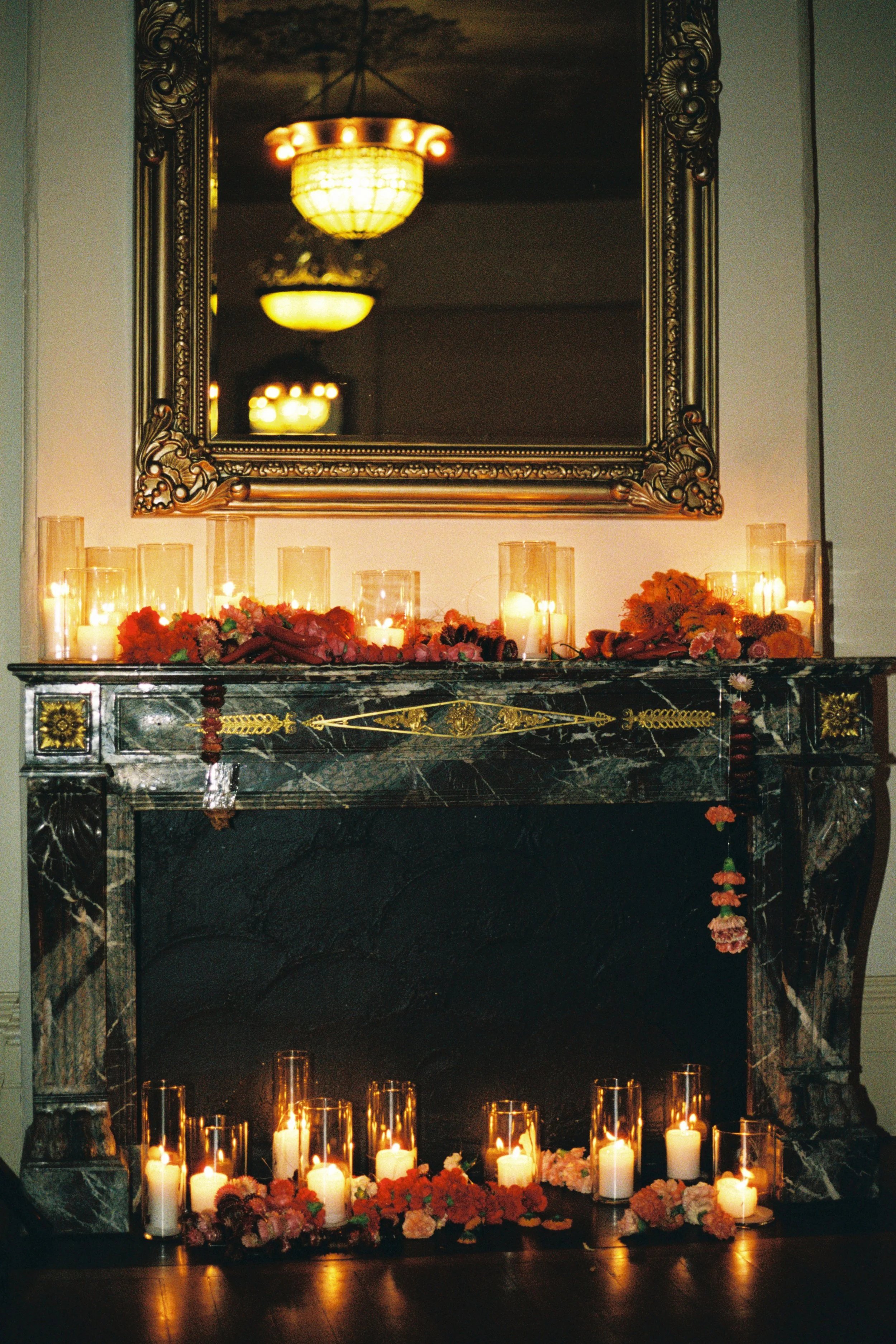 A marble fireplace decorated with lit candles in glass holders and floral arrangements, with a large ornate mirror above, creating a warm, elegant ambiance.
