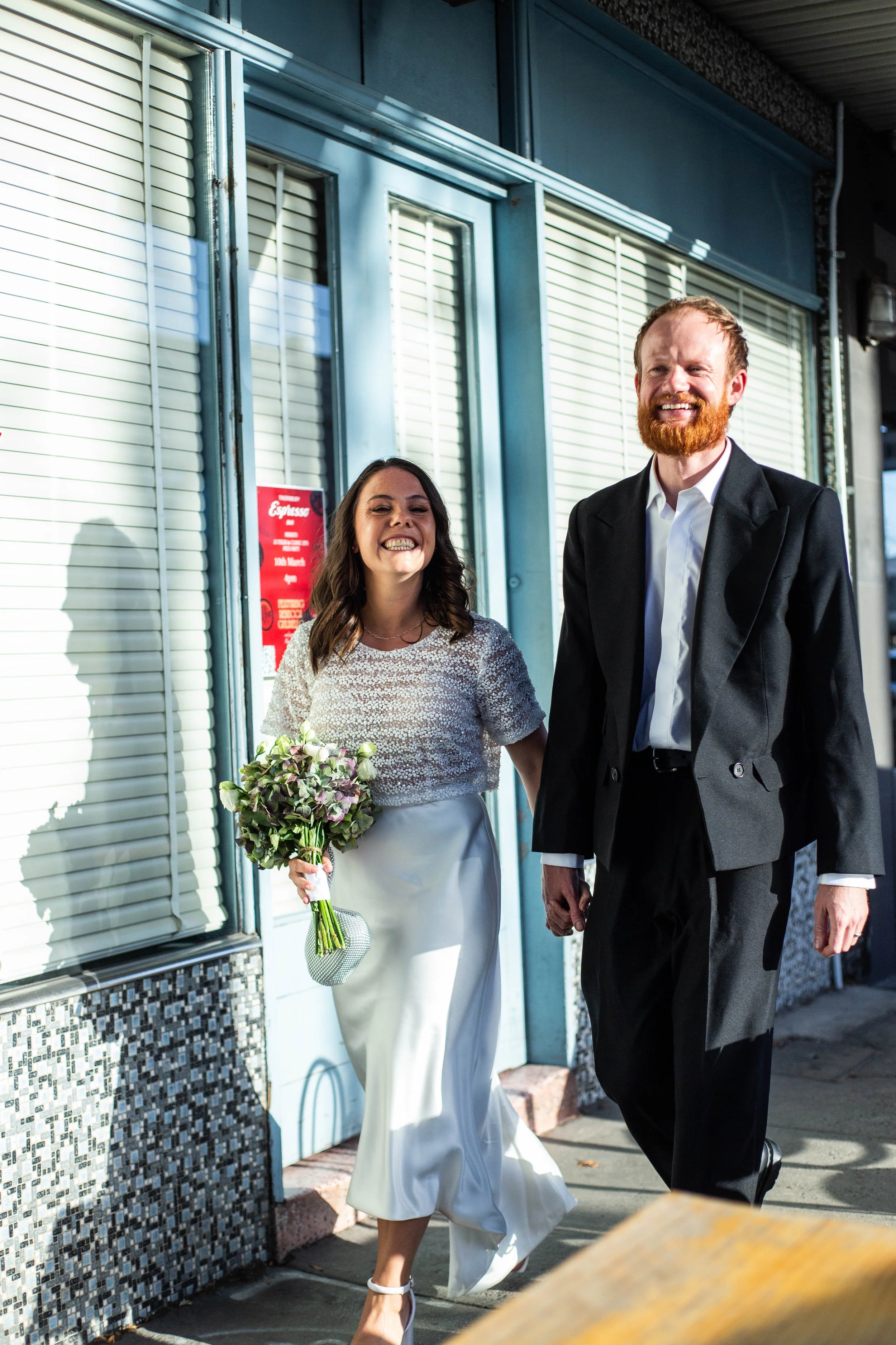 A woman in a white dress holding a bouquet of flowers walking happily alongside a man in a black suit outside a building with large windows and blinds.