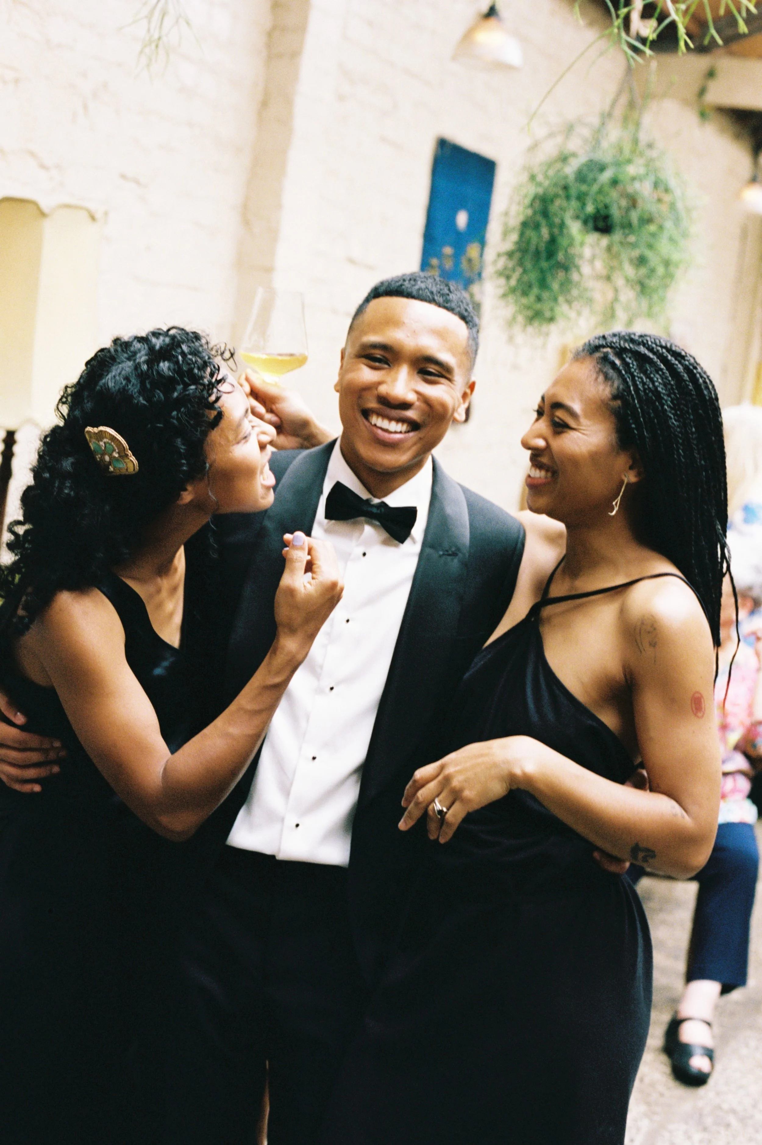 Three people celebrating at a formal event, with one man in a tuxedo and two women in elegant black dresses, smiling and holding drinks.
