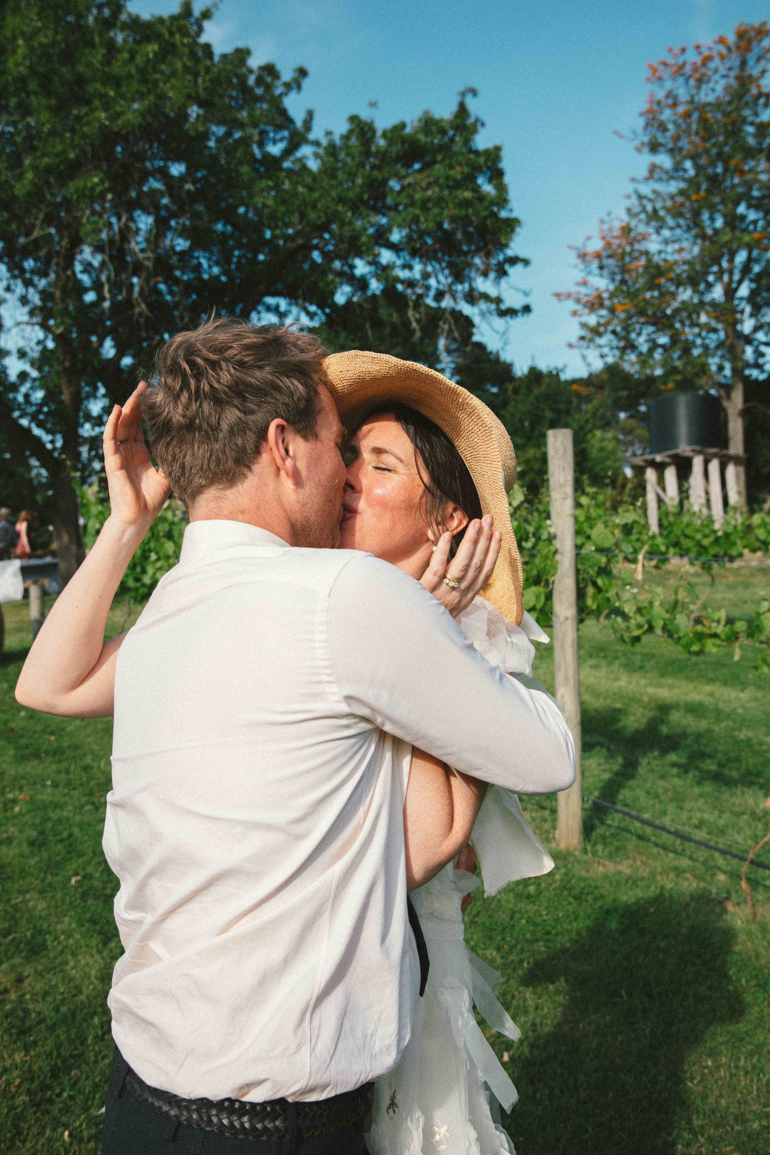 A man and a woman sharing a kiss outdoors, with the woman wearing a large sunhat and white dress, and the man in a white shirt, amidst green trees and a clear blue sky.